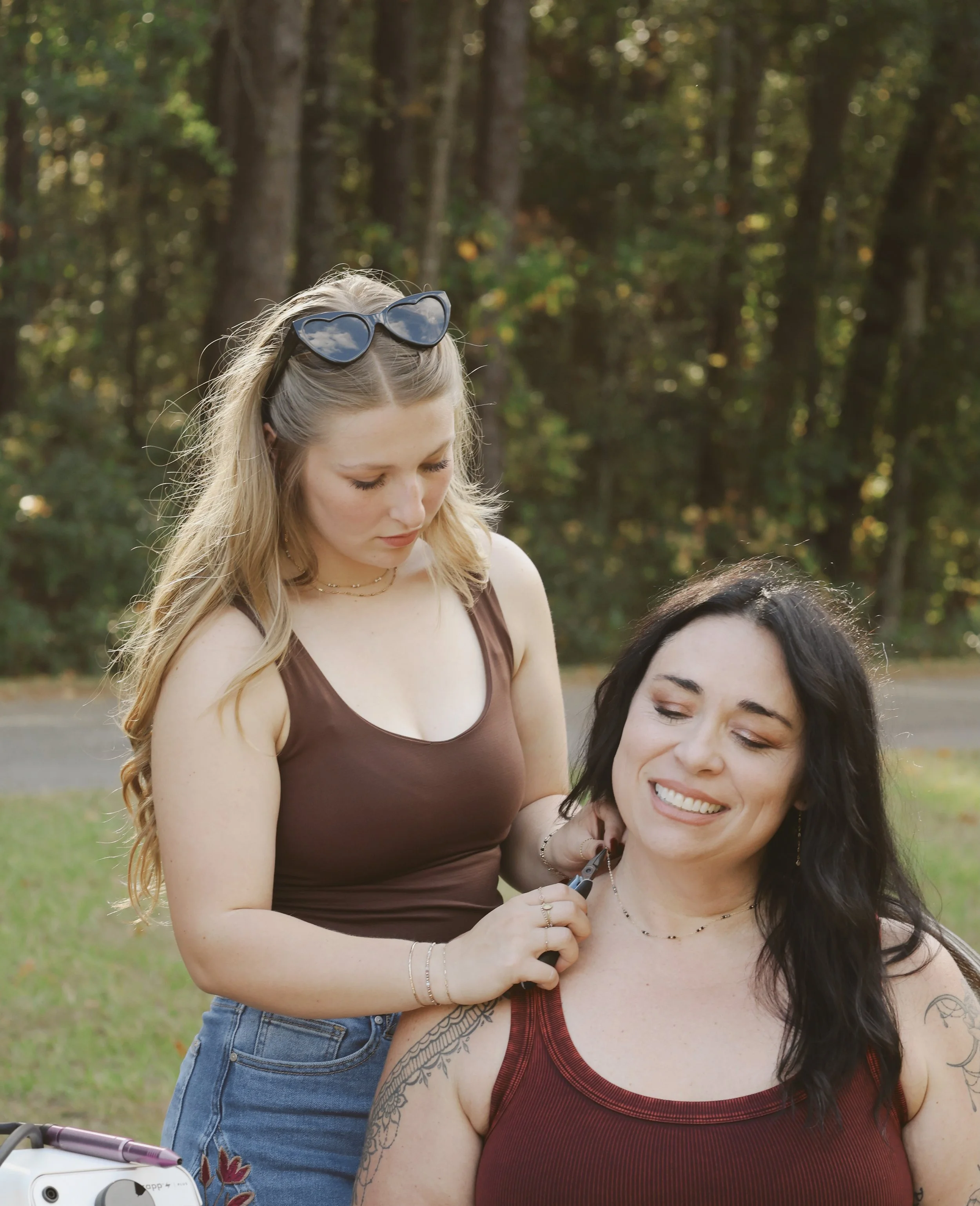Two women outdoors near a wooded area, one sitting and smiling while the other stands and is about to cut her hair.