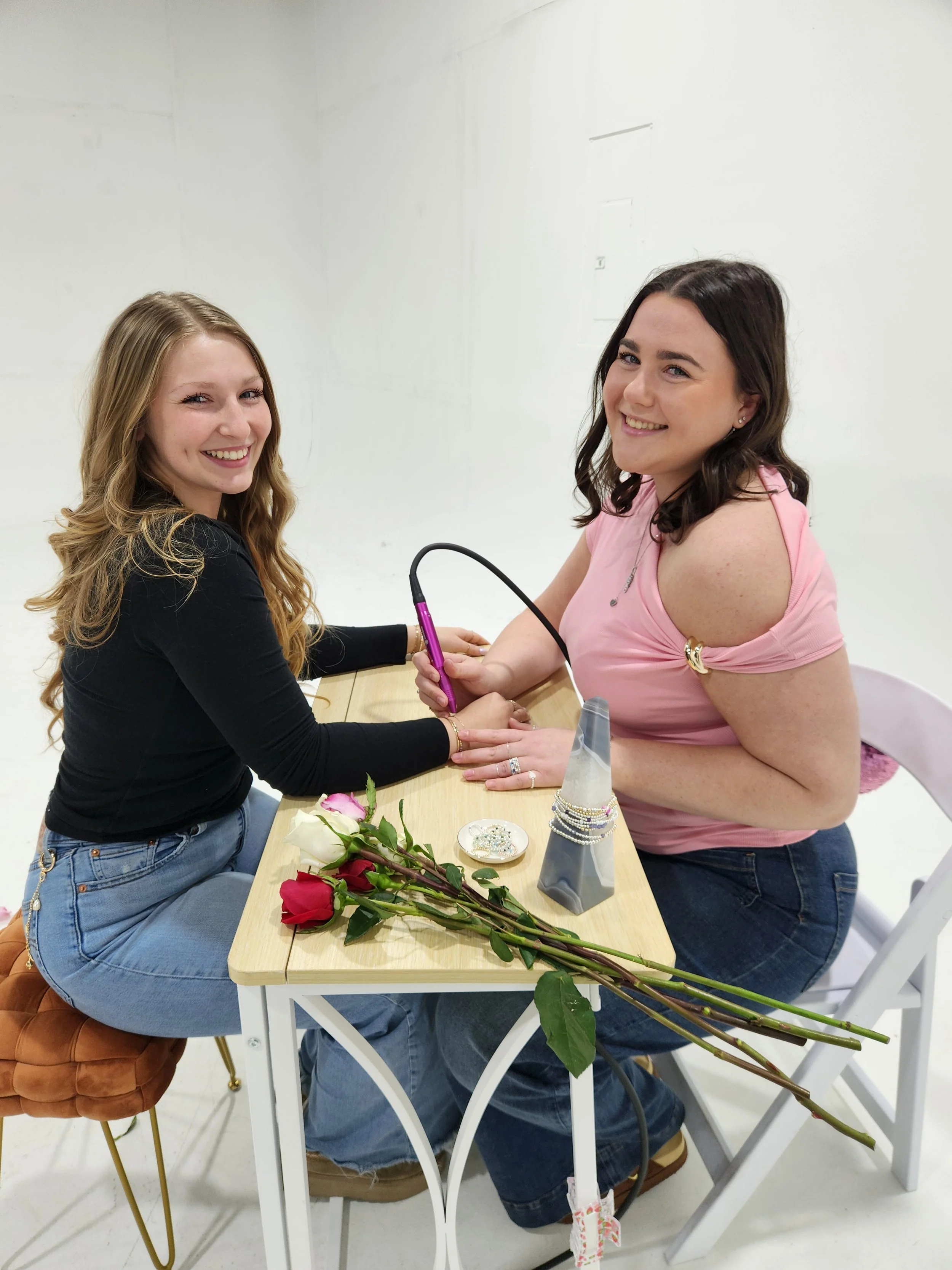Two women smiling and sitting at a table, with one woman taking a blood sample from the other. The table has three roses and some jewelry.