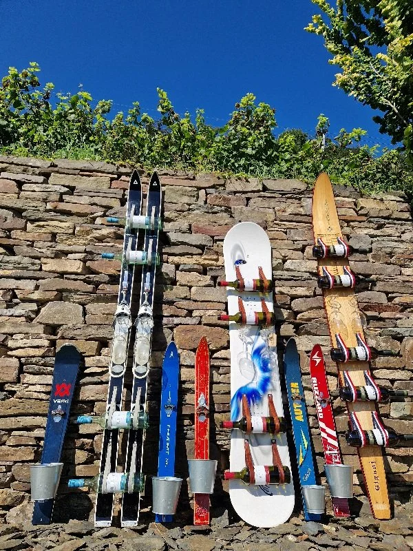 Fünf Paar Skier, die an einer Steinmauer im Freien stehen, unter einem blauen Himmel mit Bäumen im Hintergrund.