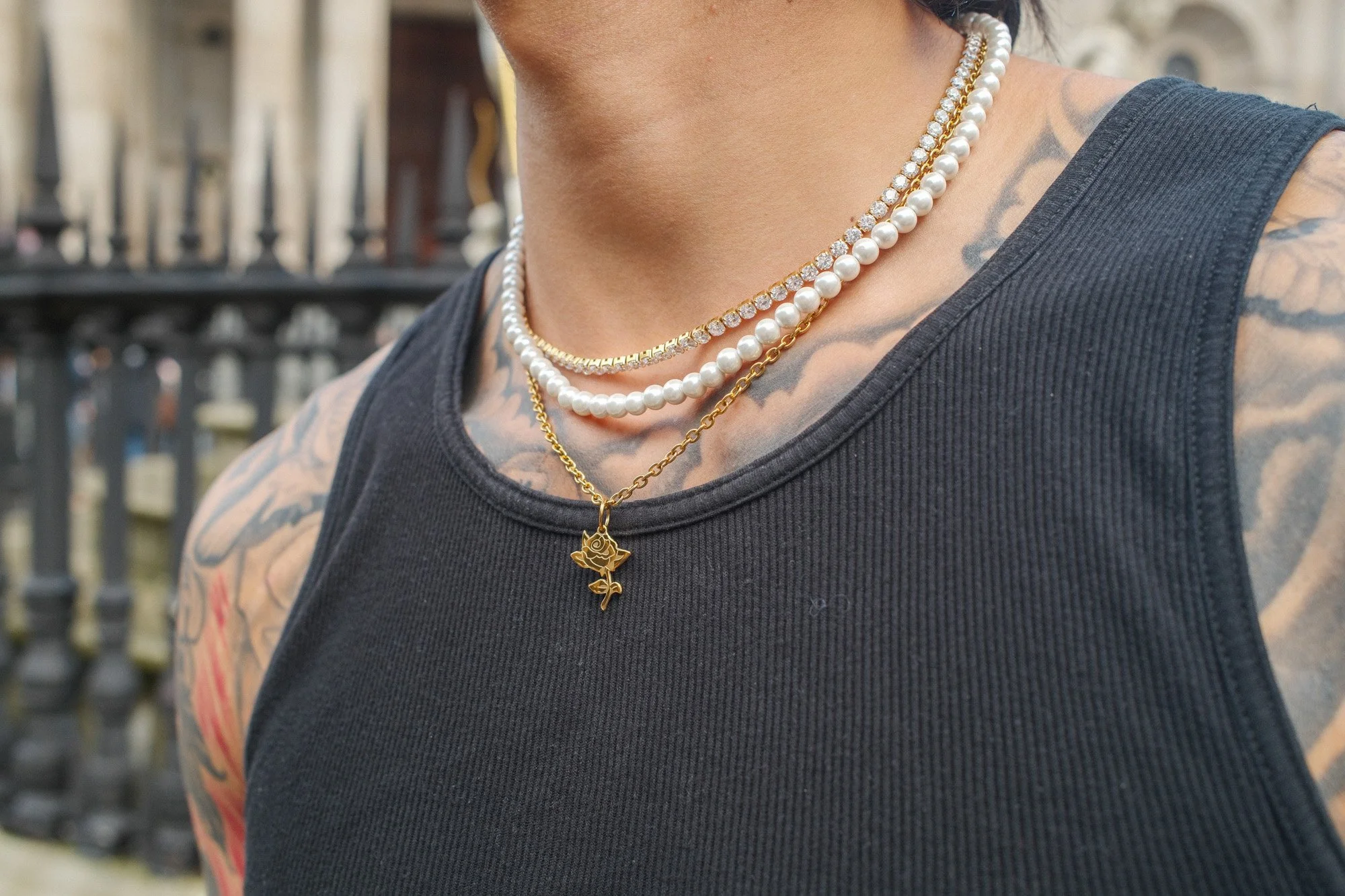 Nahaufnahme von Hals und Schultern einer Person mit Tattoos, die mehrere Halsketten trägt, darunter eine Perlenkette, eine Kette mit goldenen Elementen und eine Halskette mit einem goldenen Anhänger in Form einer Rose. Person trägt ein schwarzes ärmelloses Shirt.