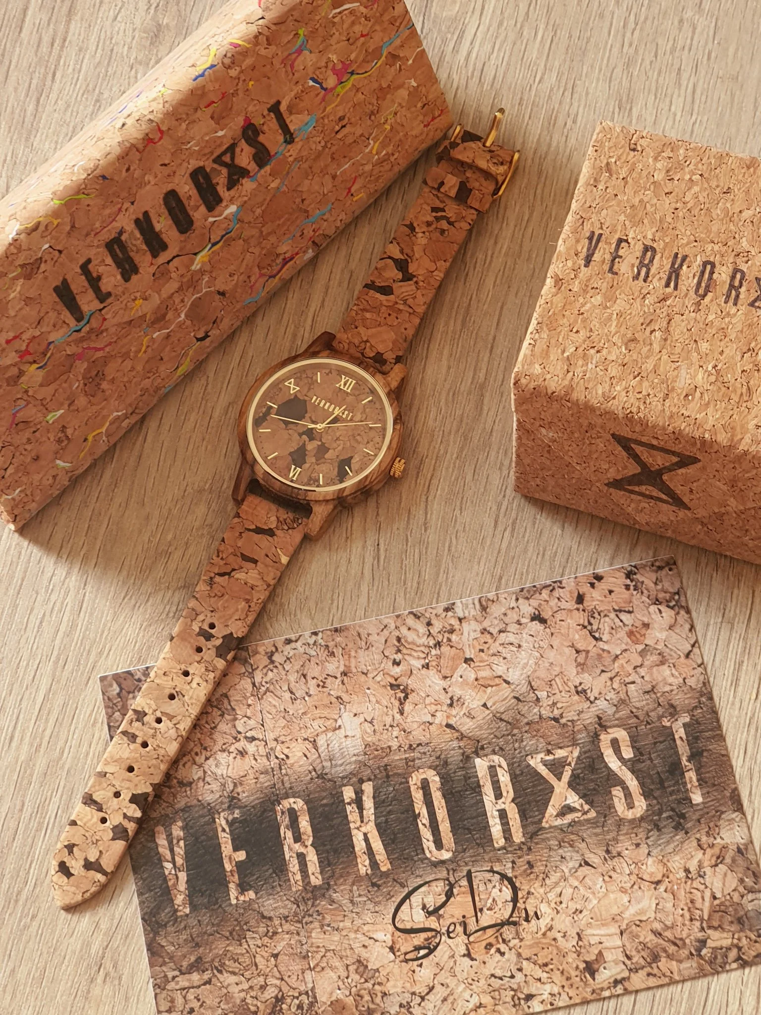 Eine Armbanduhr mit Korkband, sowie die dazugehörigen Verpackungen und ein Flyer, alles im Korkdesign. Die Uhr ist auf einem Holzoberfläche platziert.