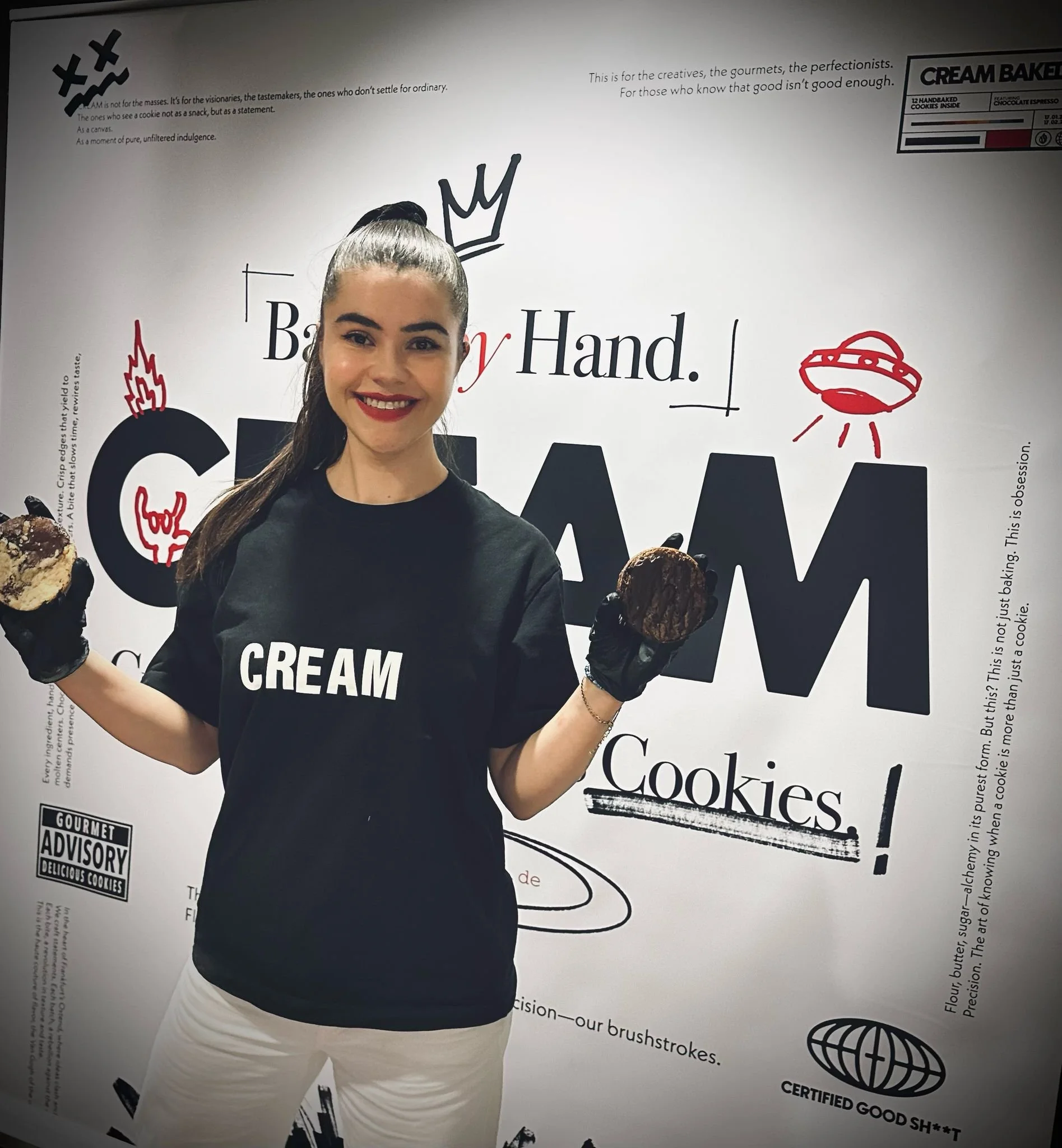 Junge Frau in schwarzer T-Shirt mit weißer Aufschrift 'CREAM' hält zwei Kekse in den Händen, vor einer weißen Wand mit verschiedenen Texten und Illustrationen rund um Kekse.