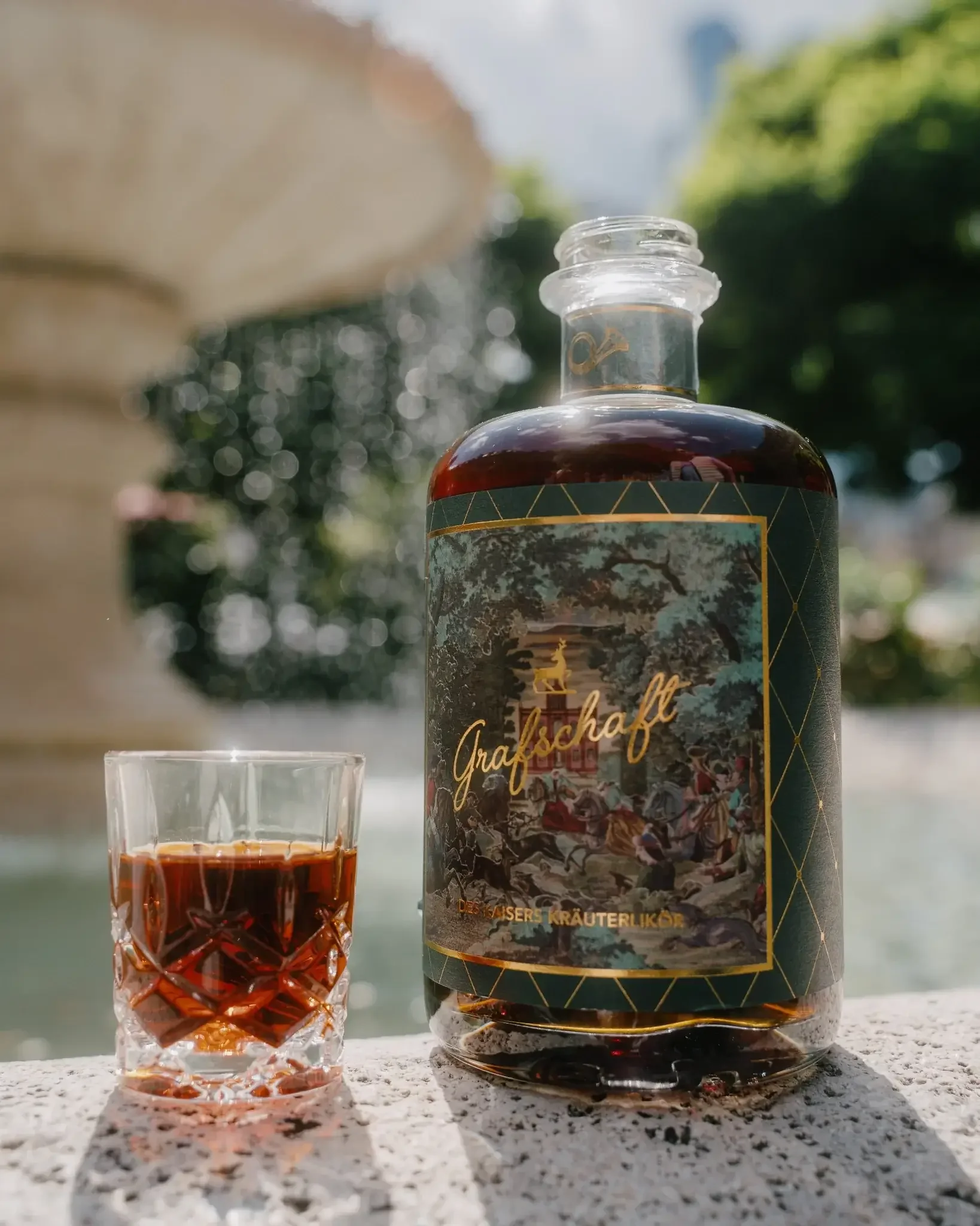 Ein Flasche Kräuterlikör mit dem Label 'Grafschaf' und einem Glas mit dunklem Likör neben einem Wasserbrunnen im Freien bei Sonnenschein.