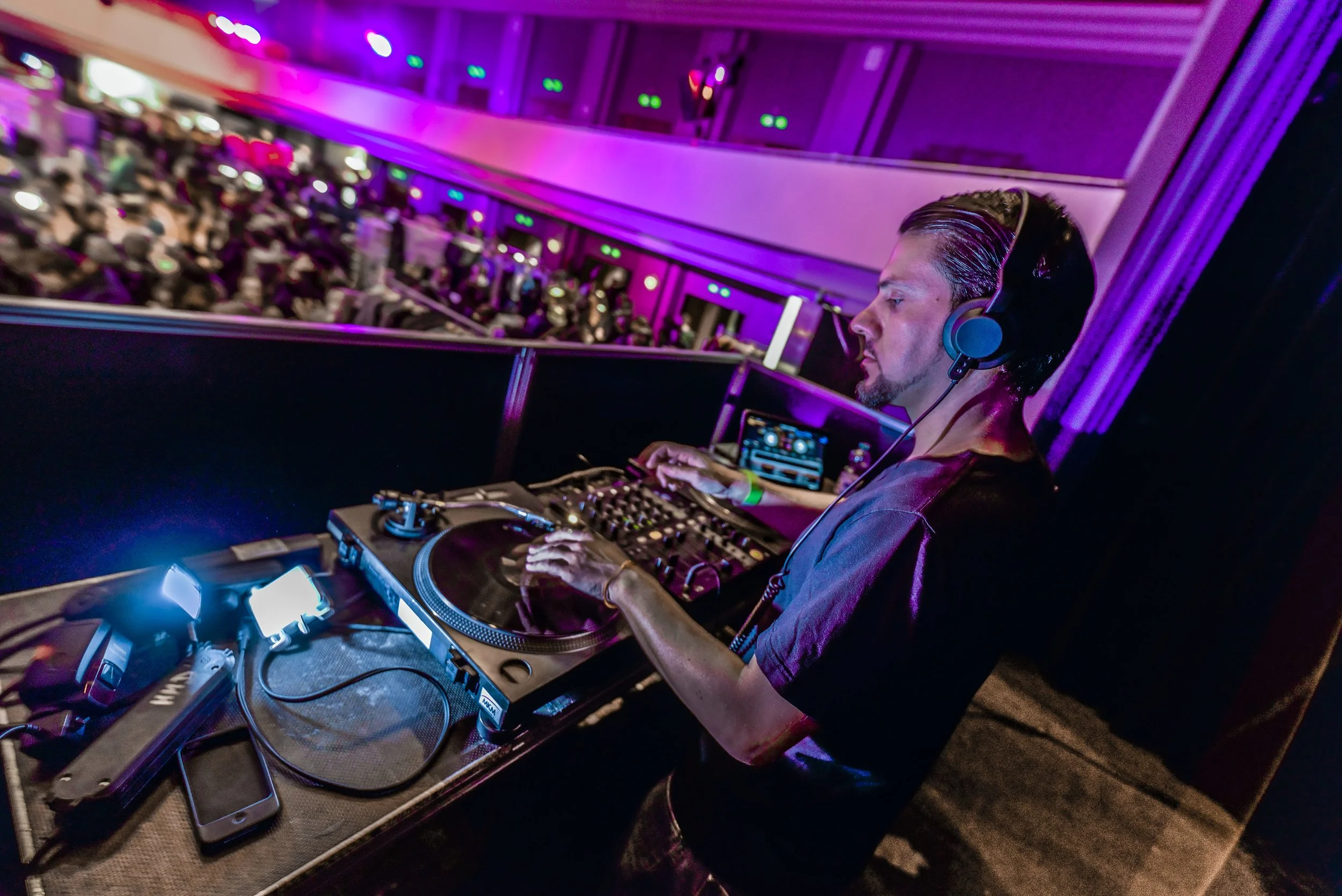 Ein DJ mit Kopfhörern an, der eine DJ-Konsole und einen Laptop bedient, während eine große Menschenmenge im Hintergrund tanzt und die Veranstaltung feiert.