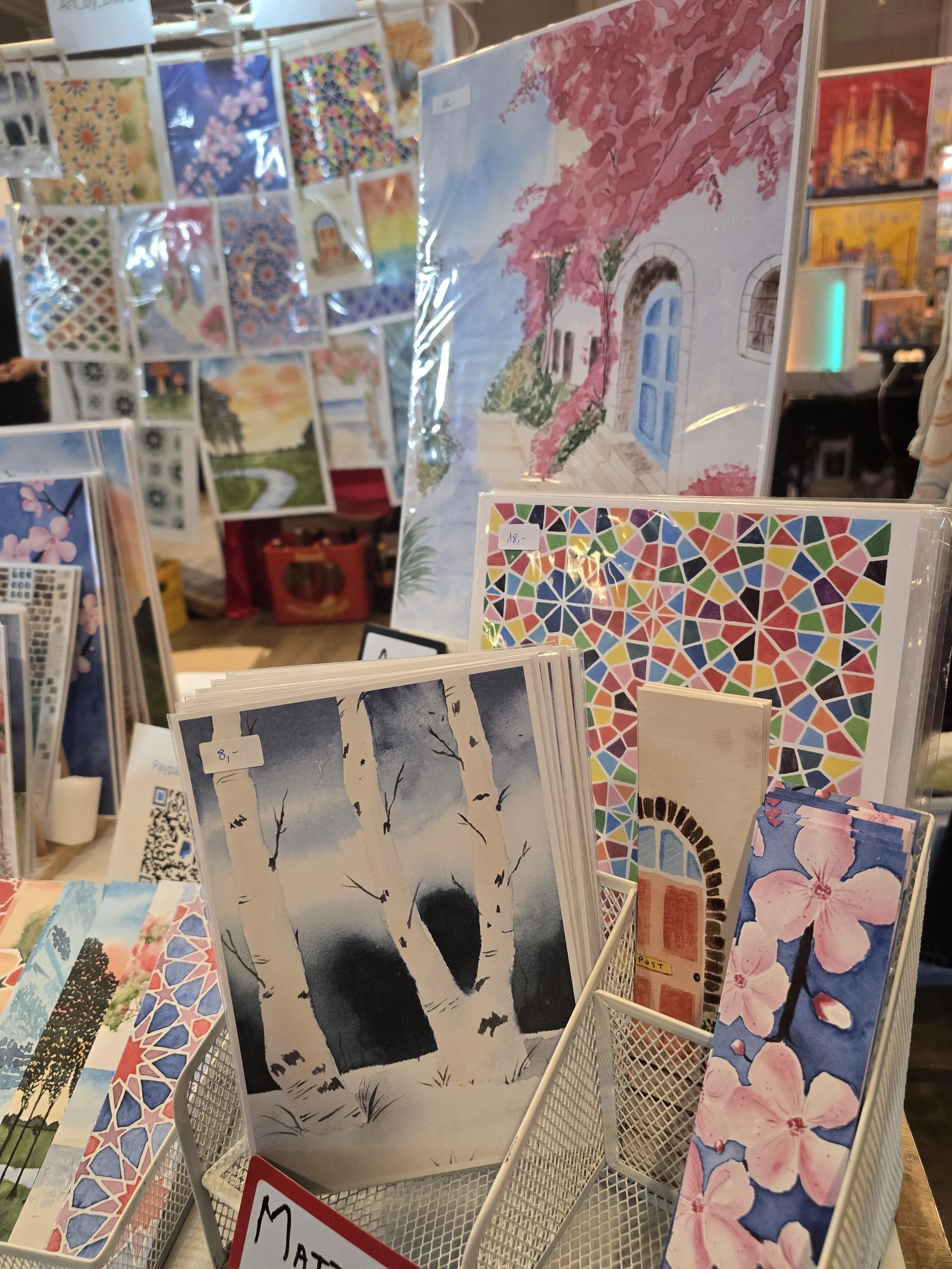 Verschiedene bunte und künstlerische Postkarten und Kunstdrucke, die auf einem Marktstand ausgestellt sind, darunter eine mit einer pink-blühenden Kletterpflanze und eine Schwarz-Weiß-Darstellung von Birken im Winter.