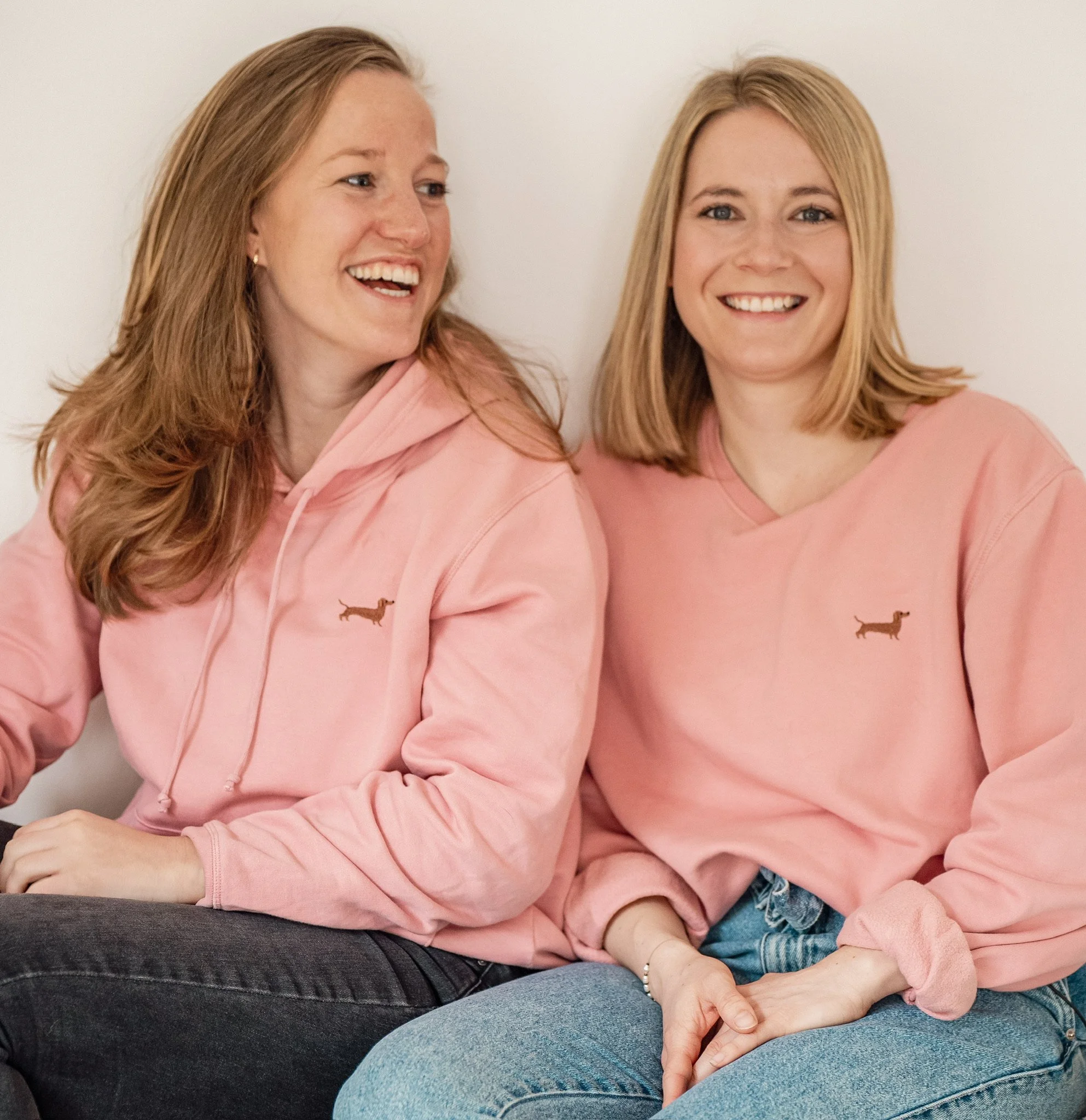 Zwei blonde Frauen sitzen nahe beieinander vor einem weißen Hintergrund, tragen beide rosa Pullover mit einem kleinen braunen Tierlogo. Sie lachen und schauen sich an.