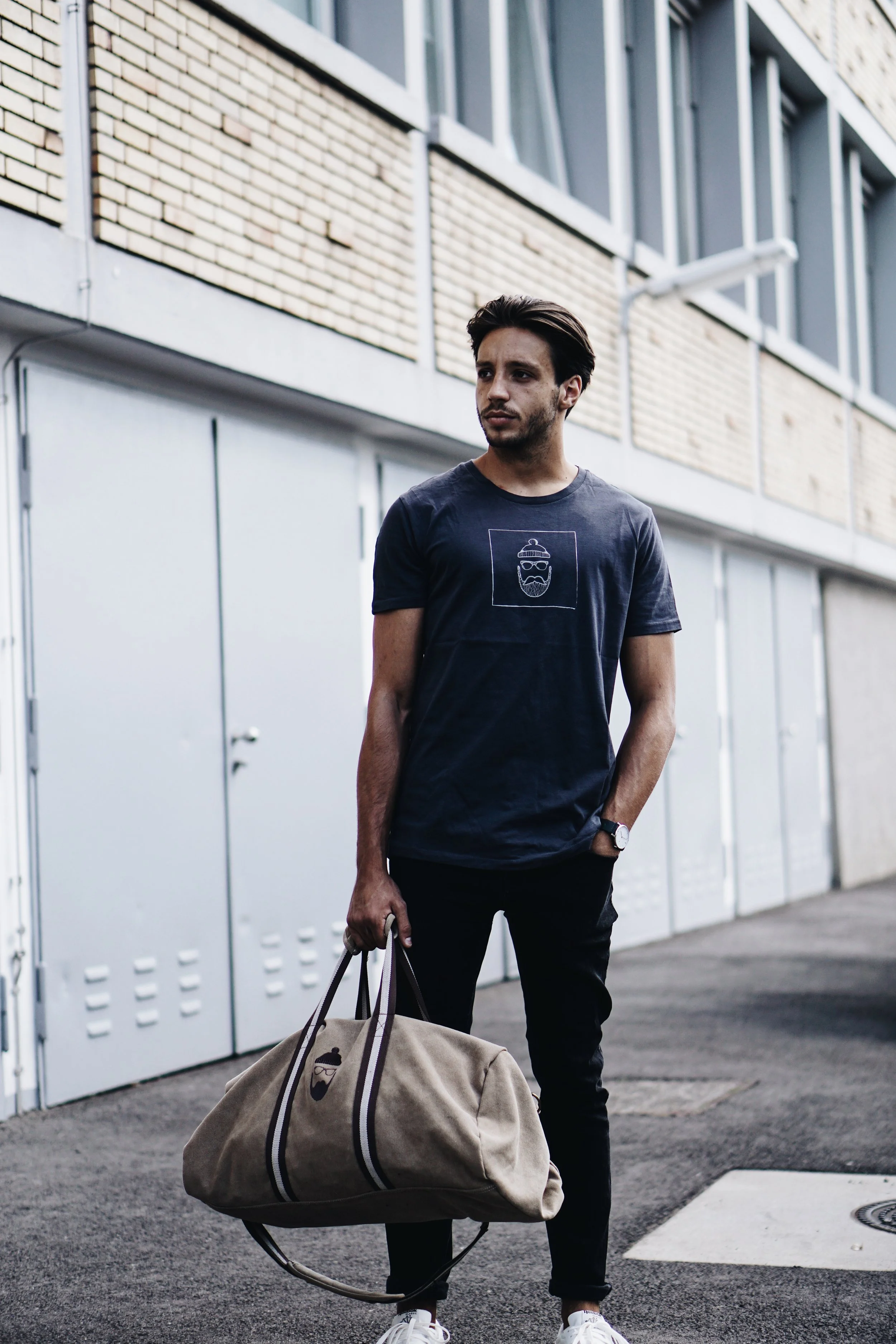 Junge Mann steht vor einer weißen Wand mit braunen Ziegelsteinen und metallic Türen, trägt ein dunkelblaues T-Shirt mit einem Logo und hält eine beige Tasche.