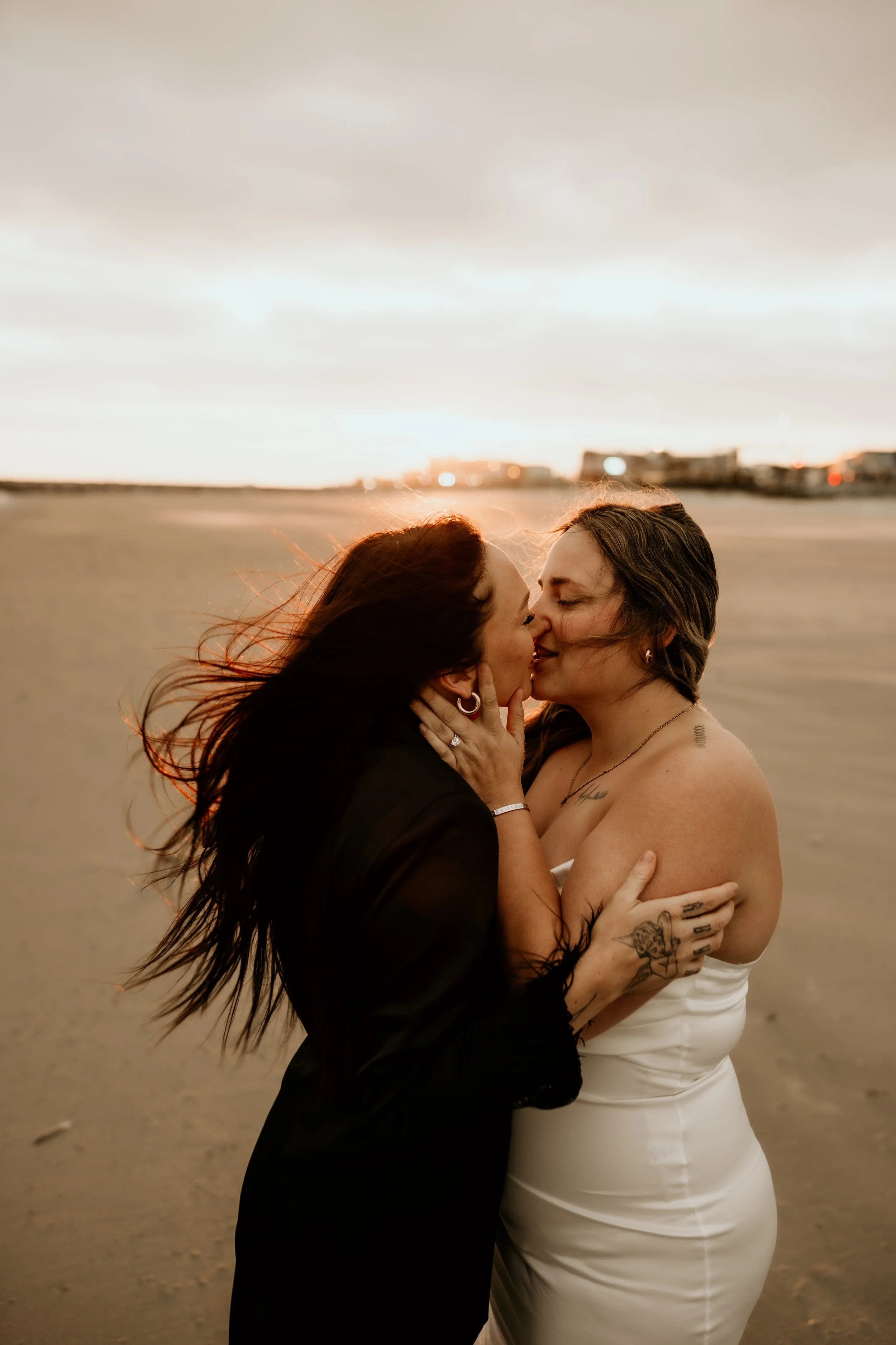 A lesbian couple on the beach in Galveston, Texas. A woman on the left wearing a black pant suit, brunette hair flowing in the wind, and a woman with a white strapless wedding dress on the right, dirty blonde hair. They are embracing and kissing.
