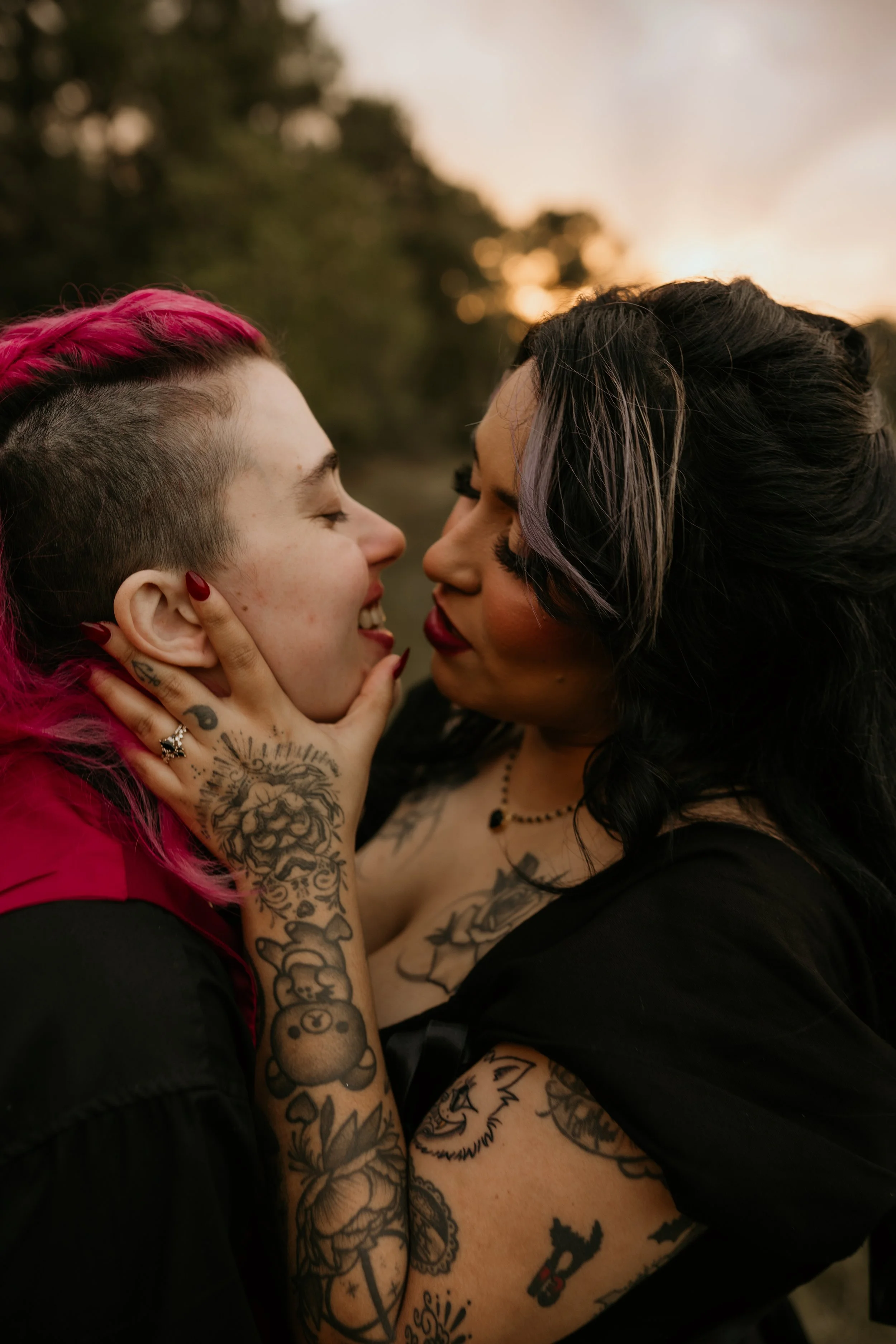 An up close of two tattooed woman kissing on their wedding day. The woman to the left has pink hair and a pink and black suit on, and the woman to the right is wearing an all black wedding dress and veil. They are kissing.
