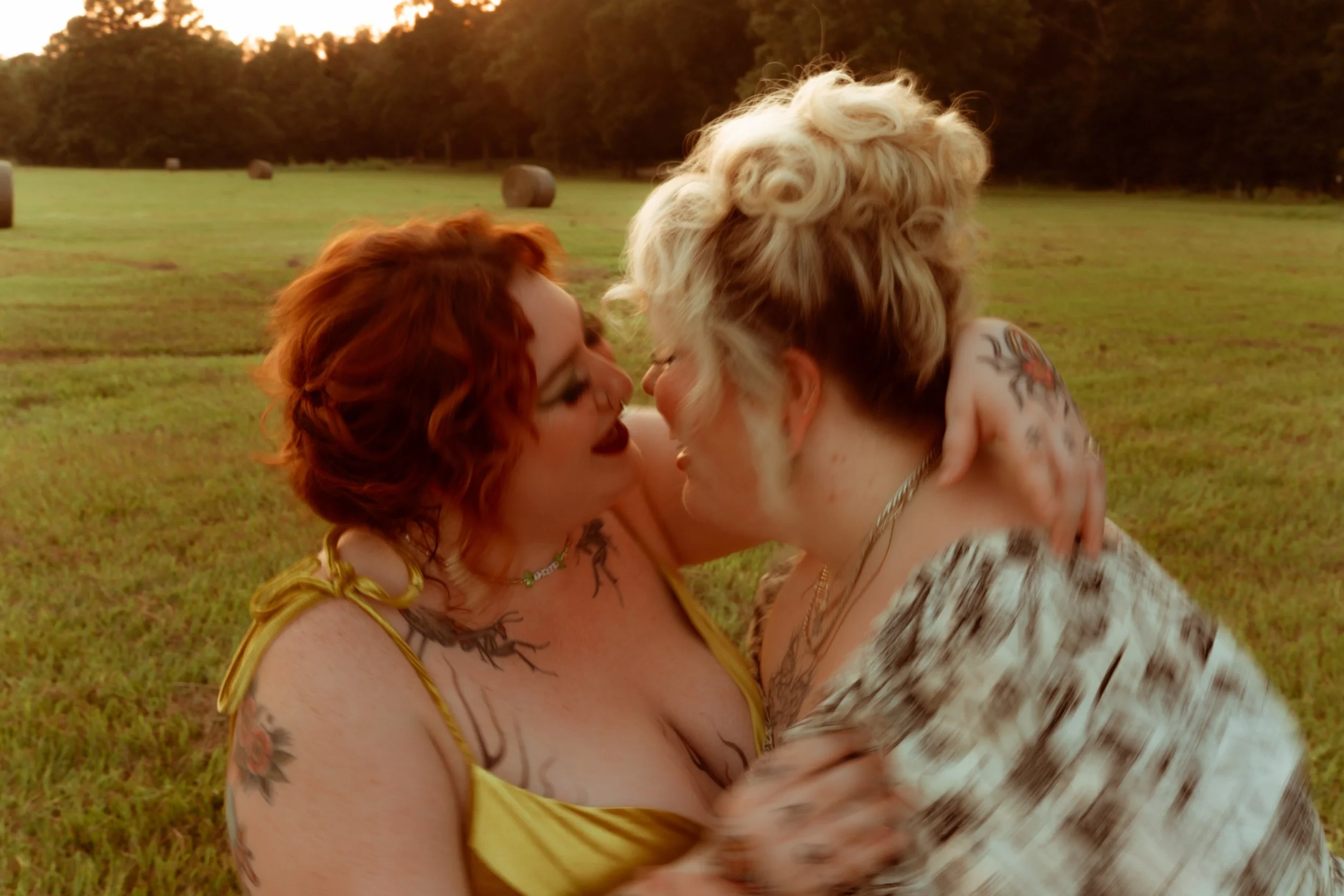 Two lesbian women embracing. The woman on the left is wearing a gold dress with thin straps, she is white and is covered in tattoos. She has red hair in a curly updo. The woman facing her is white, blonde hair also in a curly updo.