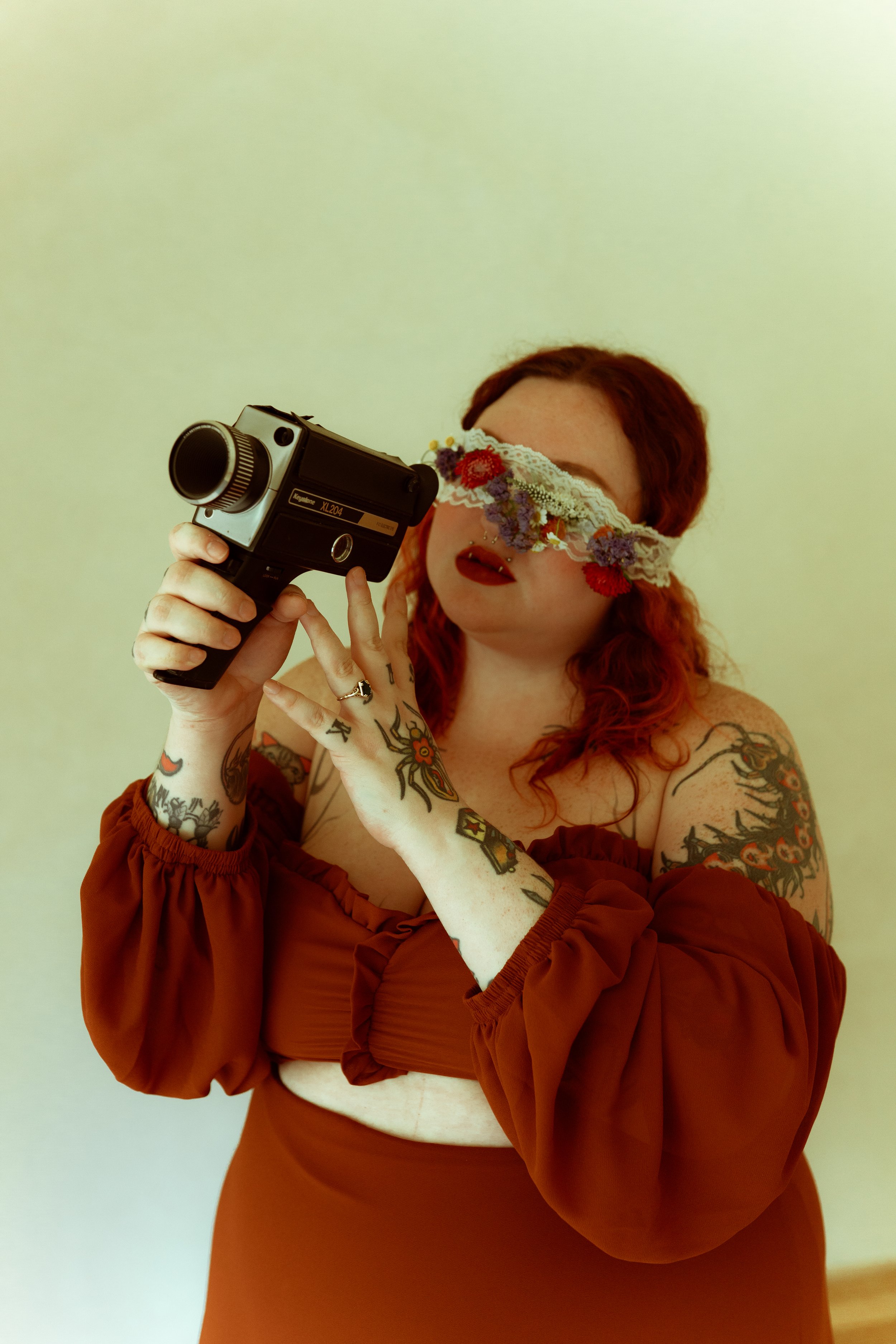 A white woman with tattoos and red hair is holding a Super 8 camera while she wears a white lace blindfold covered in flowers.