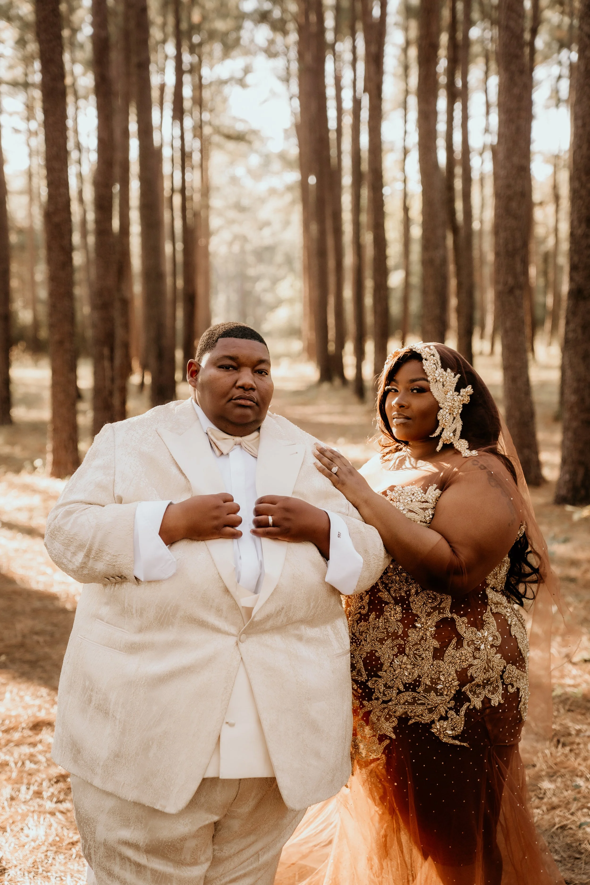 A black lesbian couple on their wedding day. The bride to the left has short brown/ black hair and is wearing an off white suit with a bow tie. The woman to the right has long brown hair with a skin toned veil and wedding dress.