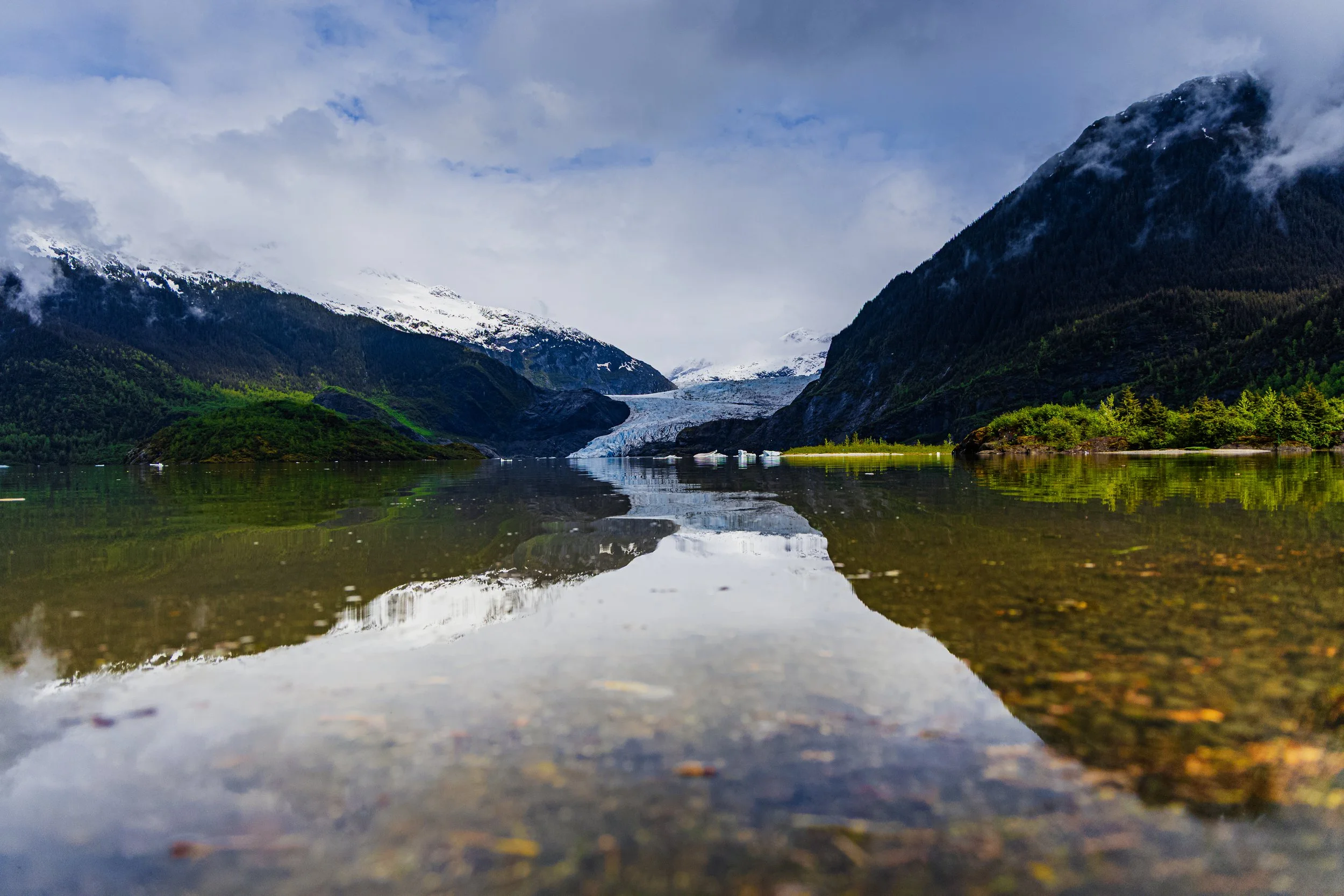 A scenic landscape with a calm lake reflecting snow-capped mountains, glaciers, and cloudy sky.