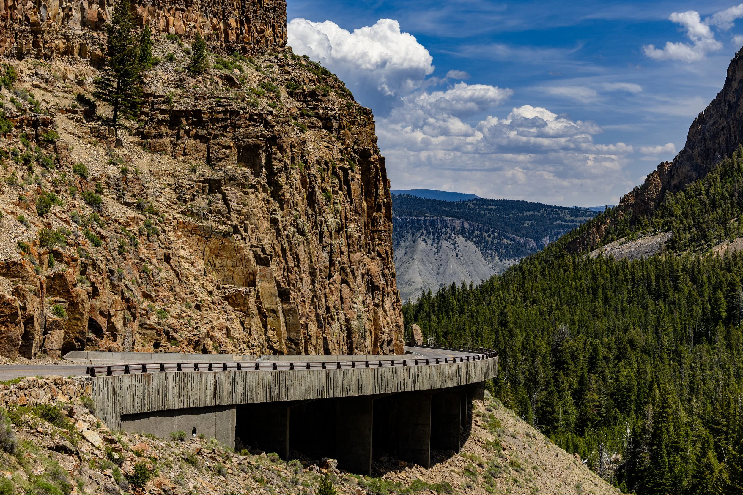 A mountainous landscape with a concrete and wooden bridge curving along the side of a rocky canyon. There are green pine trees covering the hills and a partly cloudy blue sky overhead.
