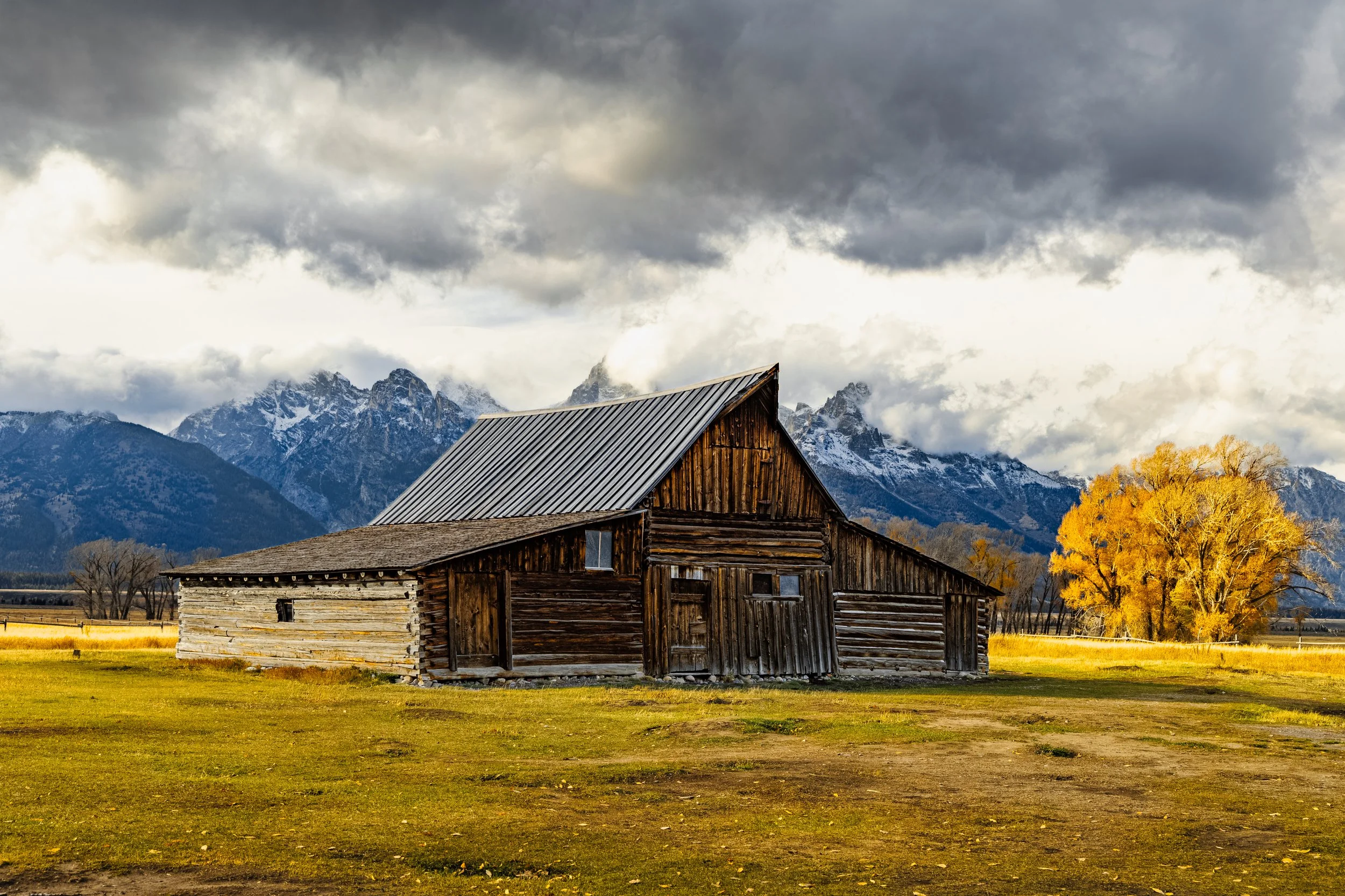 A rustic wooden barn with snow-capped mountains and cloudy sky in the background, surrounded by yellow autumn trees and open grassy fields.
