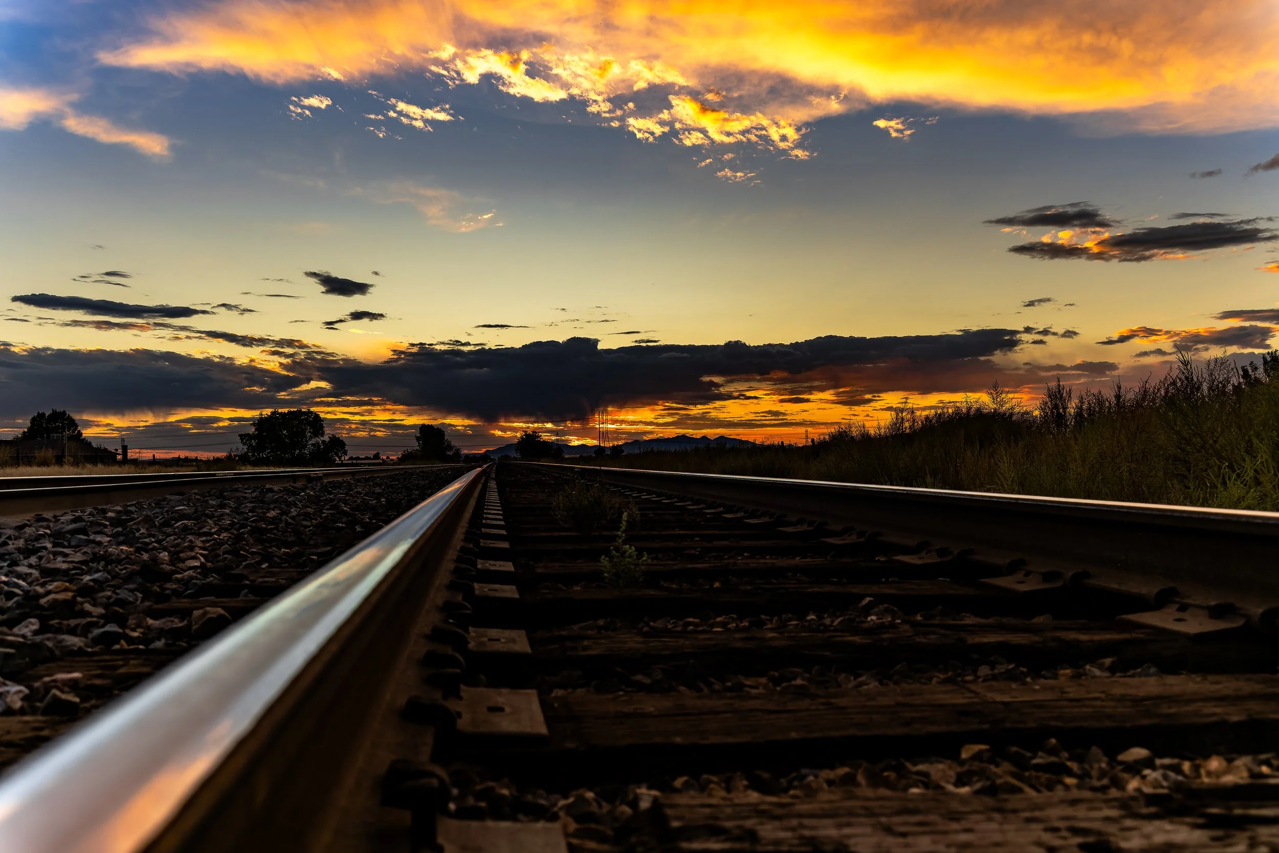 Railroad tracks extending into the horizon at sunset with a colorful sky filled with orange, yellow, and blue clouds.