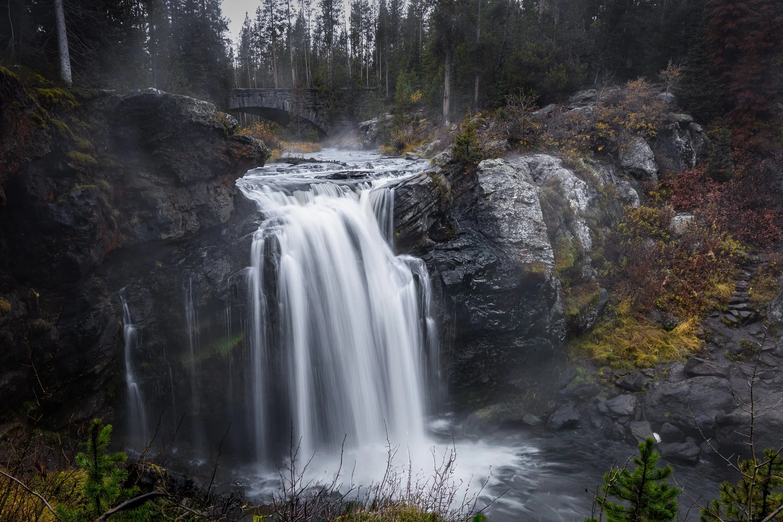 A waterfall cascading over rocks in a forested area with a stone bridge in the background.