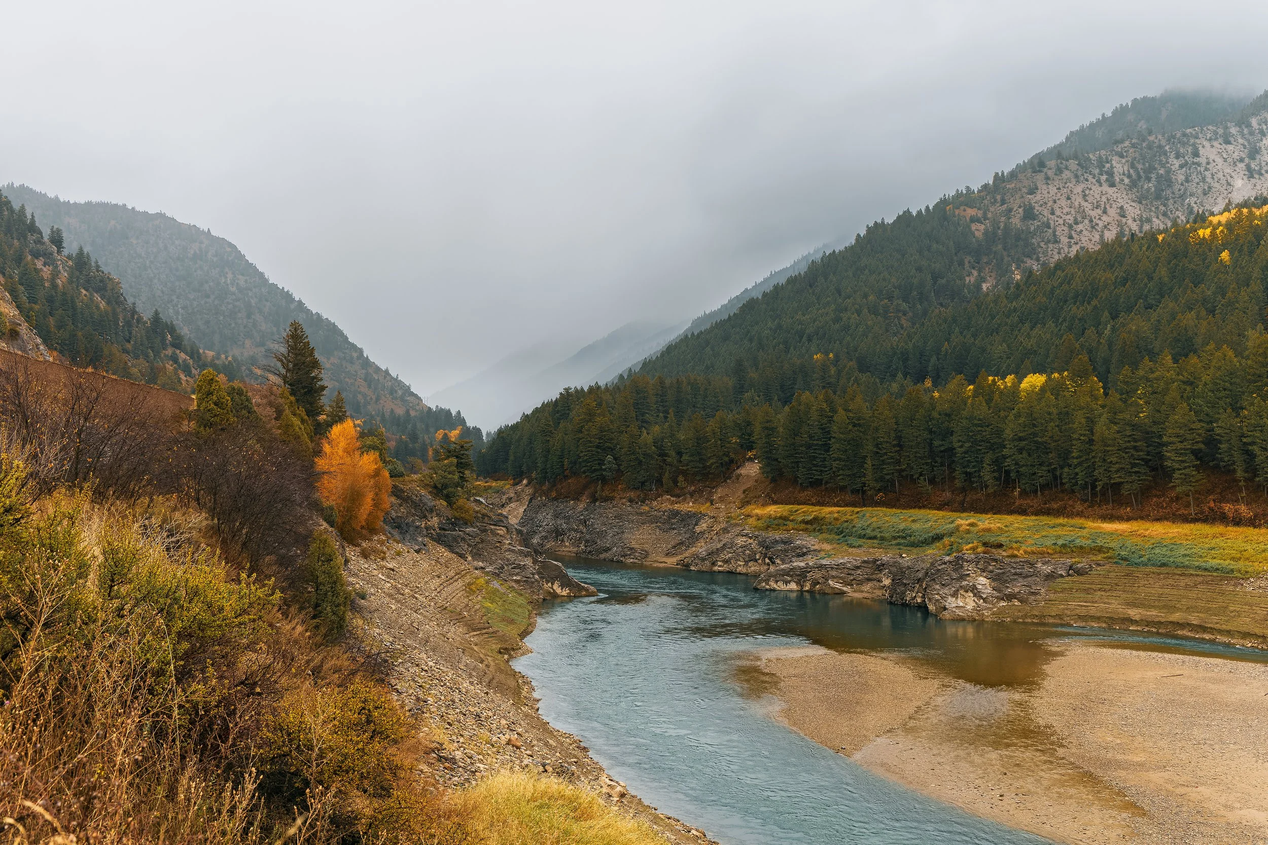 A mountainous landscape with a river flowing through the valley, surrounded by evergreen trees and colorful autumn foliage, under a cloudy sky.