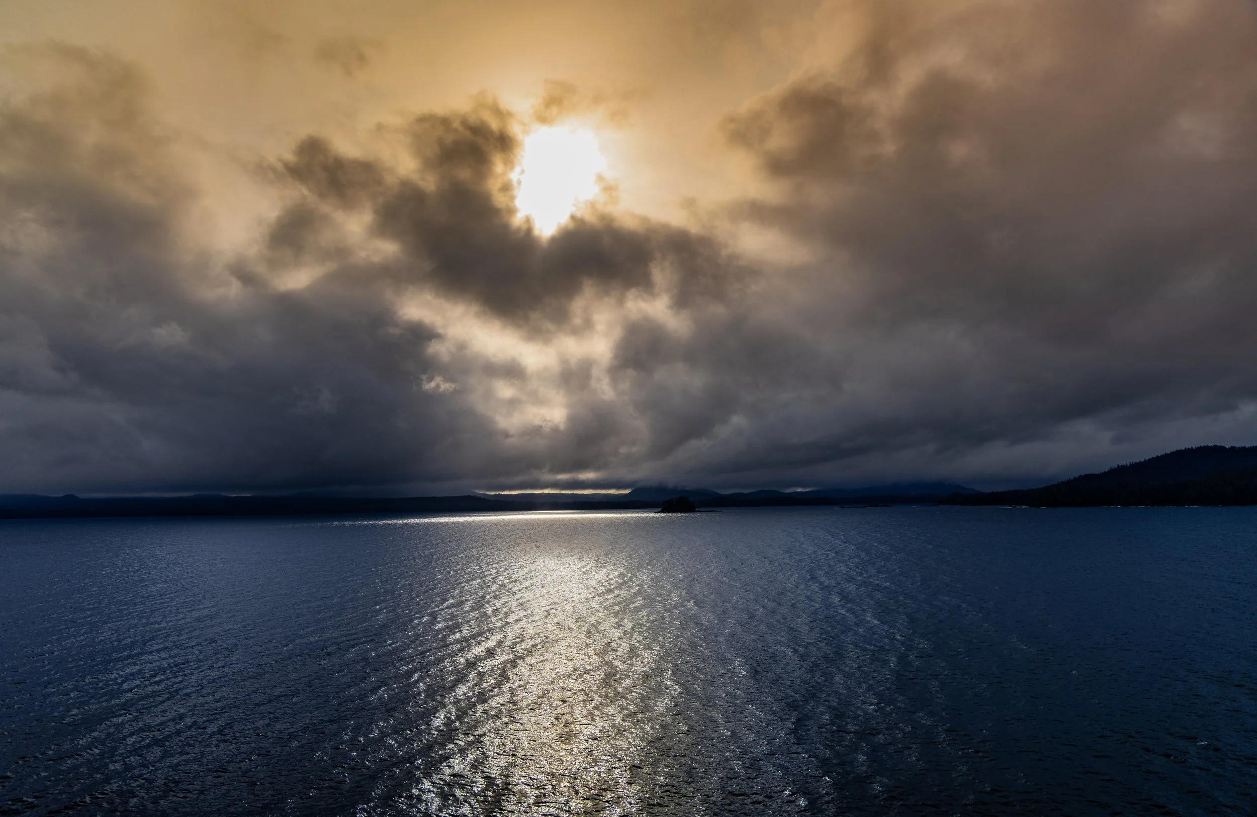 Overcast sky with the sun partially obscured by dark clouds over a large body of water, with distant hills or islands on the horizon.