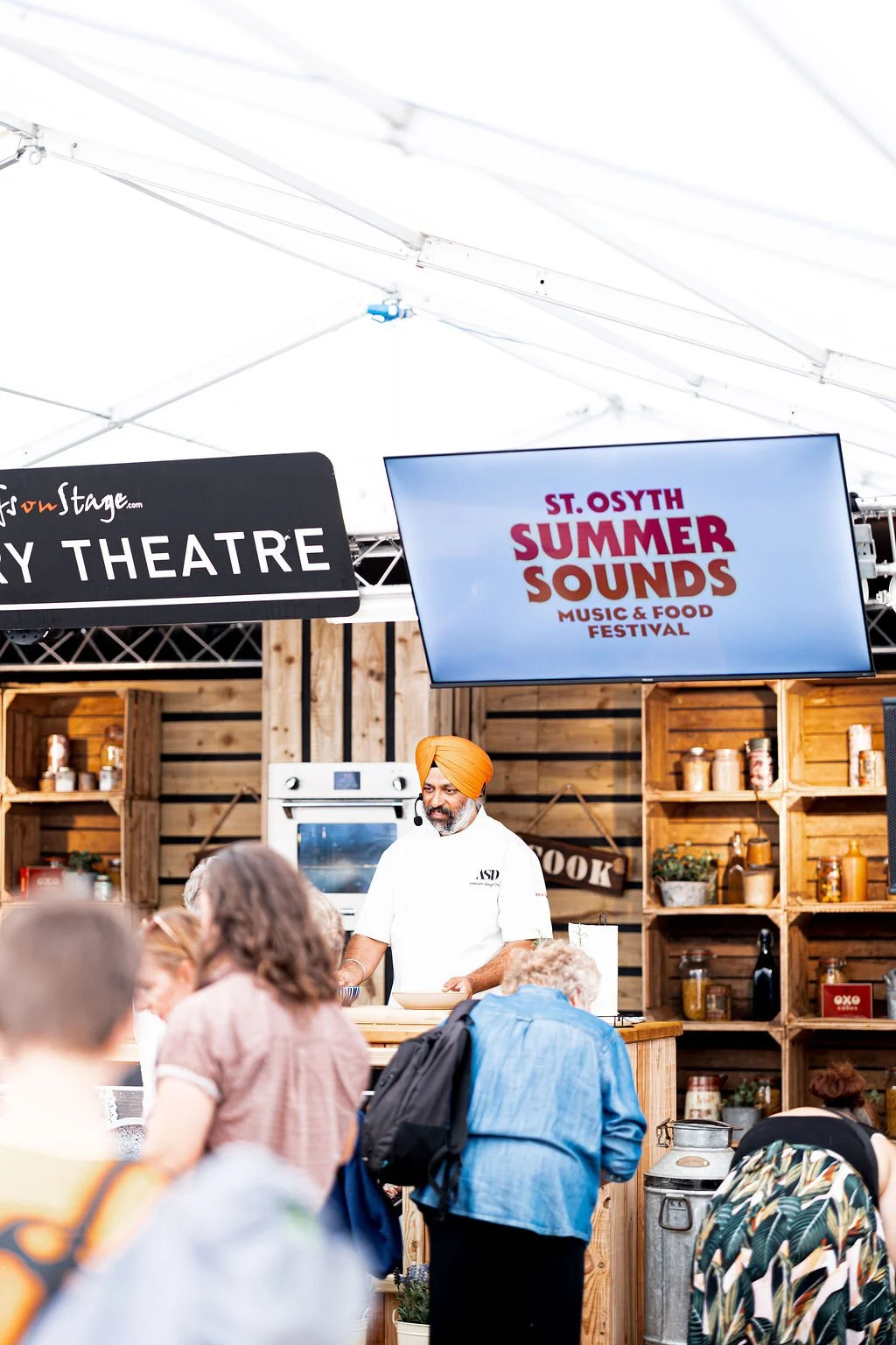 A chef wearing an orange turban and white shirt preparing food at a stall during St. Osyth Summer Sounds music and food festival. The background features wooden shelves with jars and bottles, and a large screen displaying the festival's name and details.