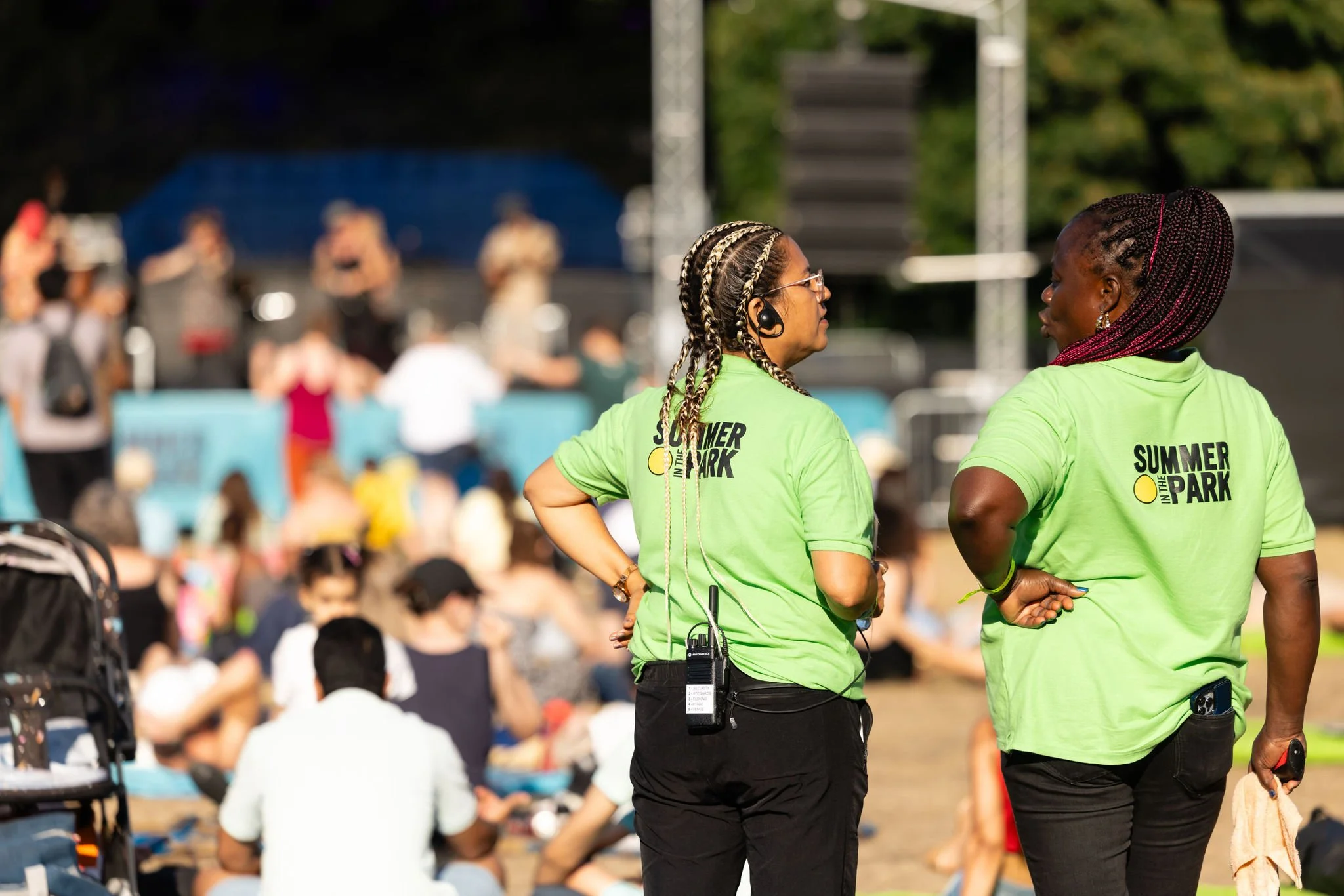 Two women wearing green 'Summer in the Park' shirts standing and talking at an outdoor event with a crowd of people seated and a stage in the background.