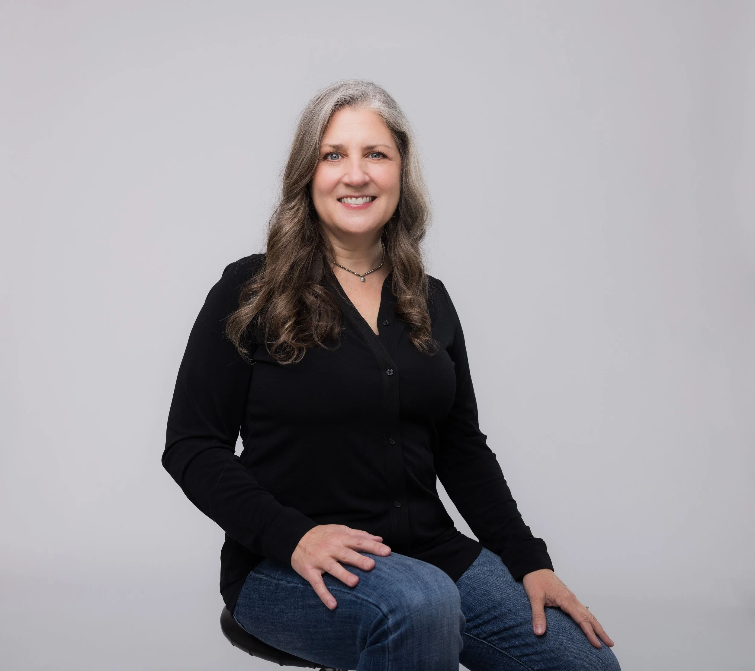 Portrait of a woman with long, wavy gray hair, wearing a black button-up shirt and blue jeans, sitting on a stool against a plain gray background.