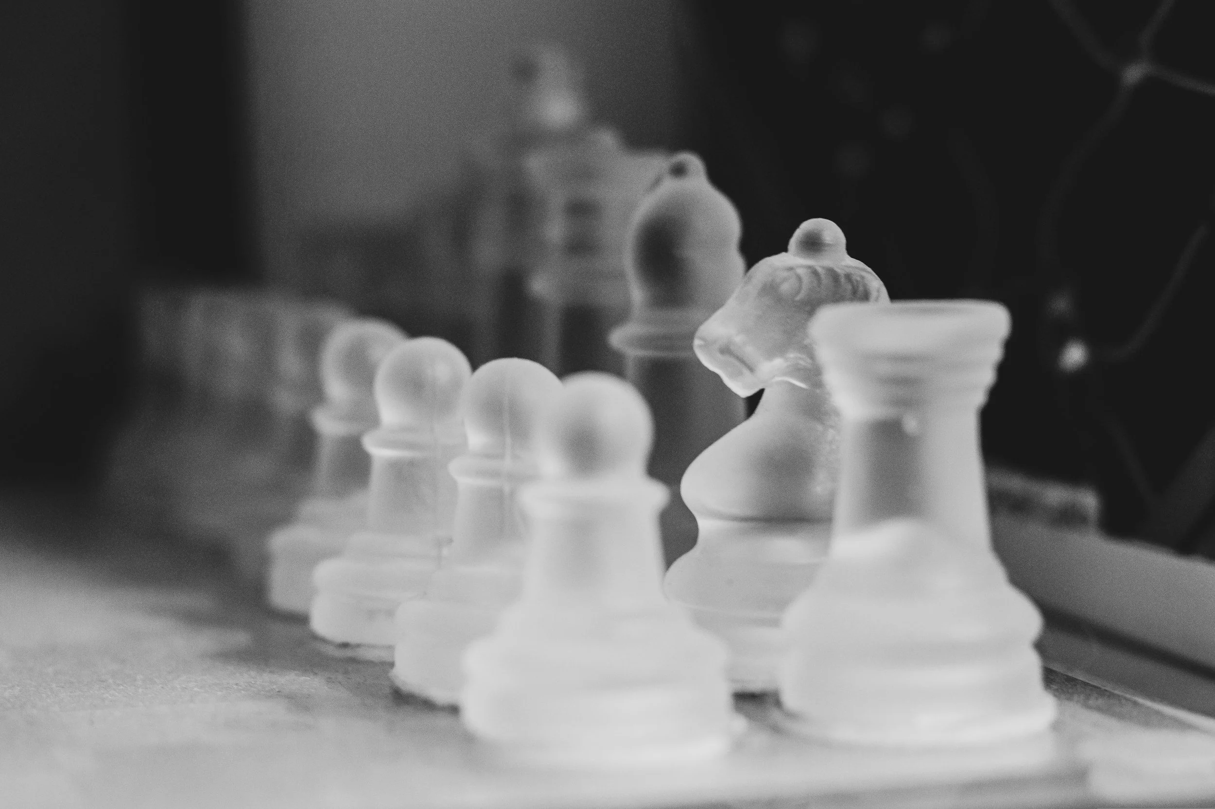 Black and white photo of glass chess pieces arranged on a chessboard, with a focus on the bishop and a pawn in the foreground and other pieces blurred in the background.