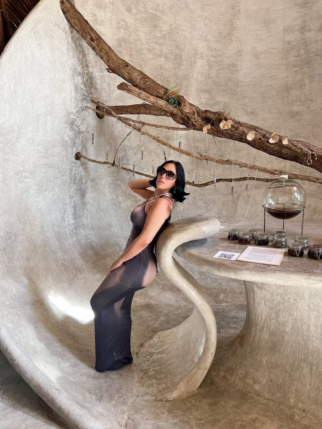 A woman in a black dress and sunglasses posing inside a uniquely shaped concrete structure with a curved bench and table, a large glass container filled with dark liquid, and a branch suspended from the ceiling with hanging objects.