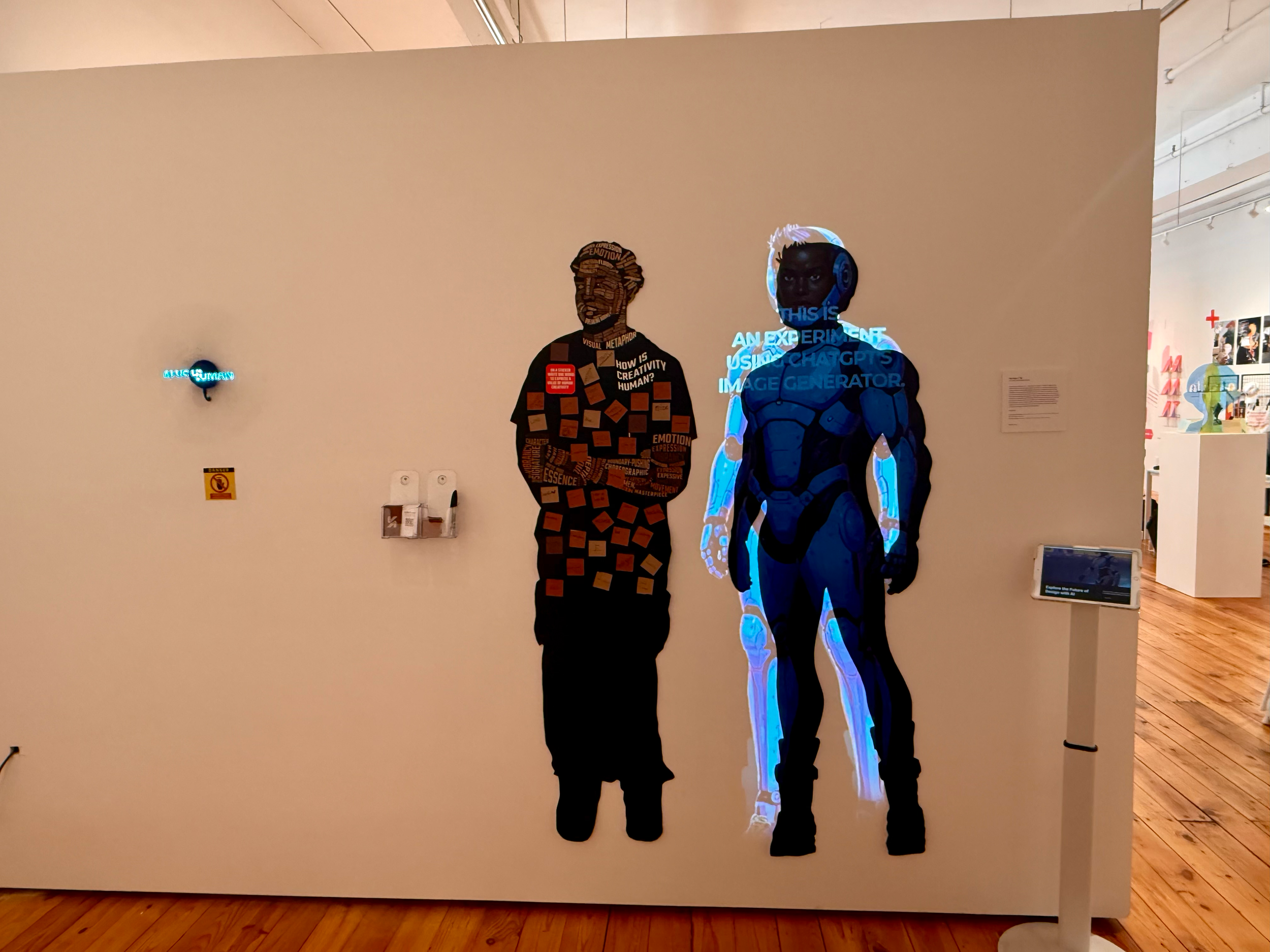 An exhibit featuring a cutout of a person with a human face and a robotic figure with electronic features, illuminated with blue text and lights in a gallery setting.