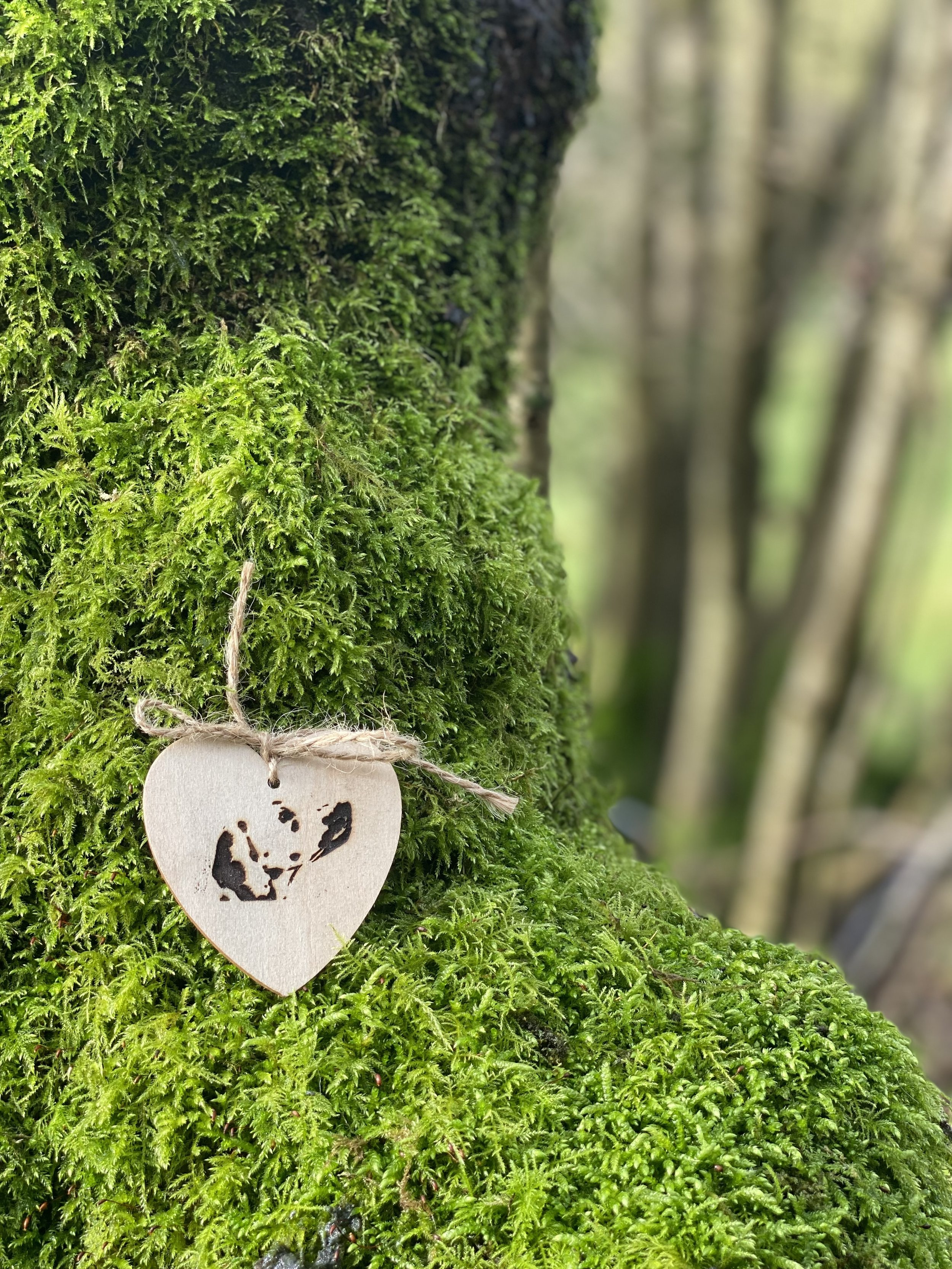 A small wooden heart ornament with a black silhouette of a dog, hanging on a green mossy tree trunk by a twine bow in a forest setting. A pet cremation keepsake