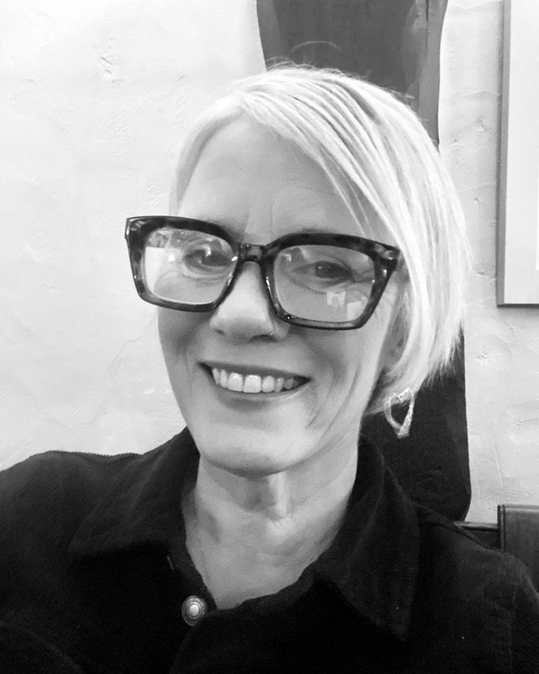 Black and white photo of a woman, Natasha of Pet Crematorium in South Wales with short blond hair, wearing large glasses and earrings, smiling at the camera.