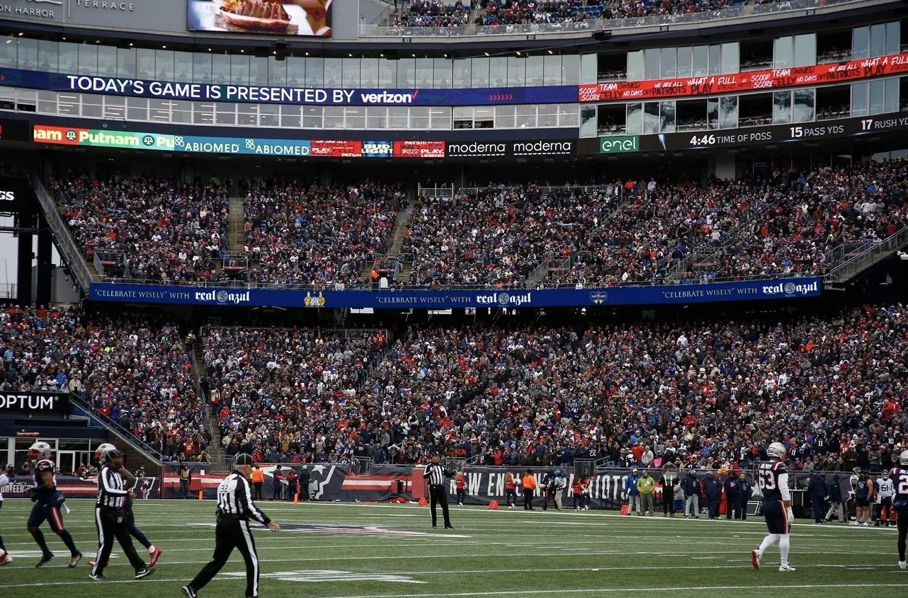 A football game at a stadium filled with spectators, with players on the field and digital advertising banners around the stands.