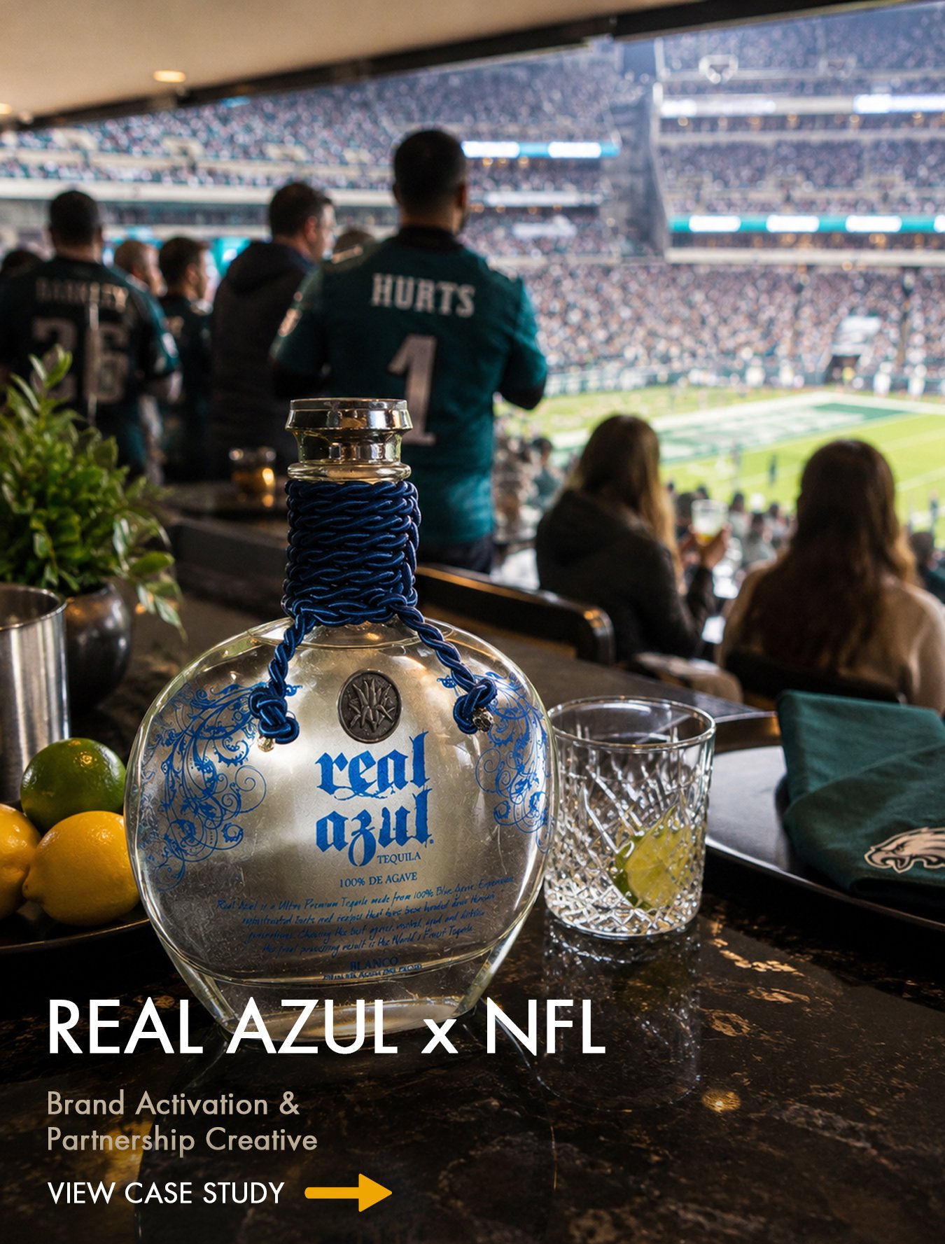 A bar at a football stadium with a bottle of Real Azul tequila on the counter, surrounded by lemons and a glass of lemon water, with spectators watching a game in the background.