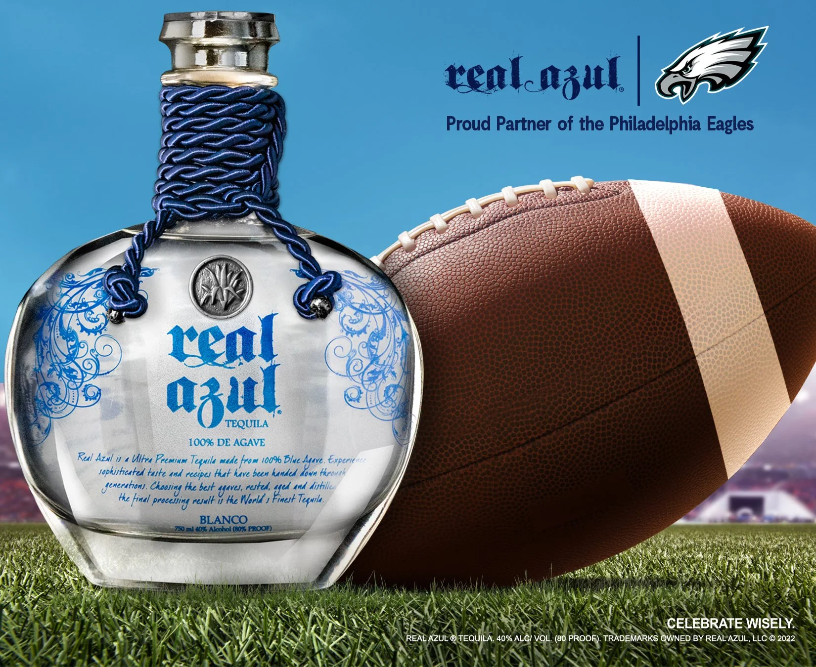 A bottle of Real Azul tequila with a blue braided cord around the neck, placed on a green football field with a brown and white football beside it. The background is clear blue sky.