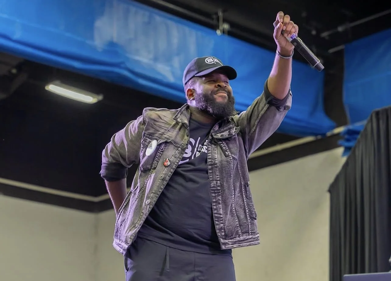 A man with a beard and cap holding a microphone and smiling at an event.