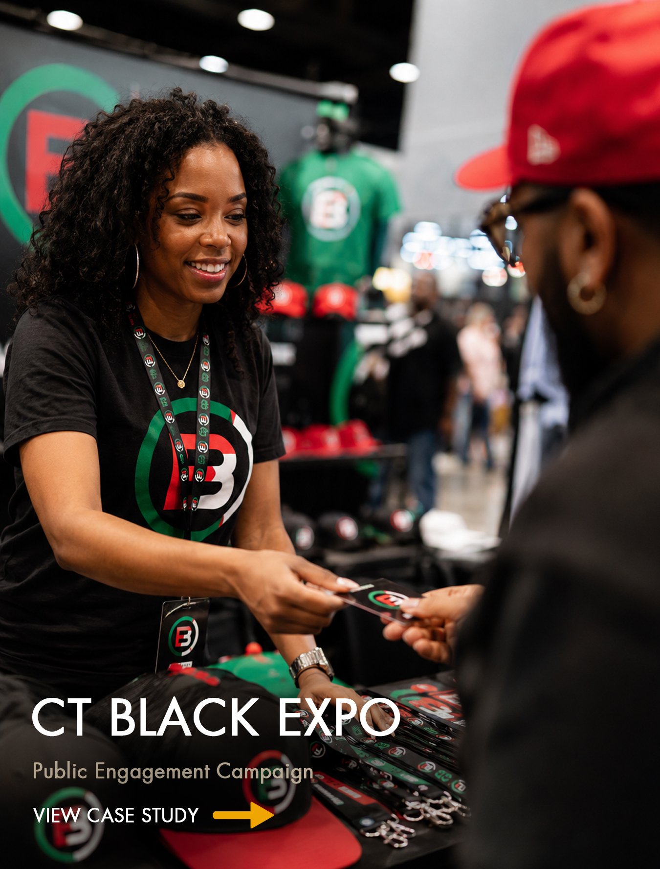 A woman with curly hair smiling and handing a card to a man at a booth with merchandise and a large sign that reads 'CT BLACK EXPO.' The booth displays hats, keychains, and other items featuring a logo with a red, black, and green design.