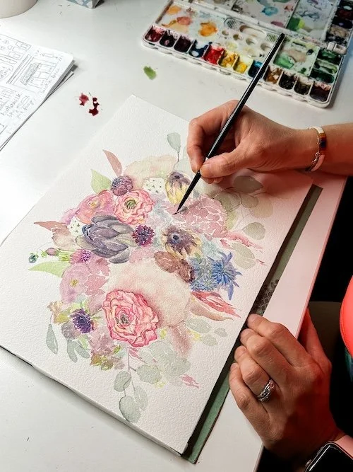 Person painting a floral watercolor design, holding a brush over a palette of colors, on a table with art supplies.