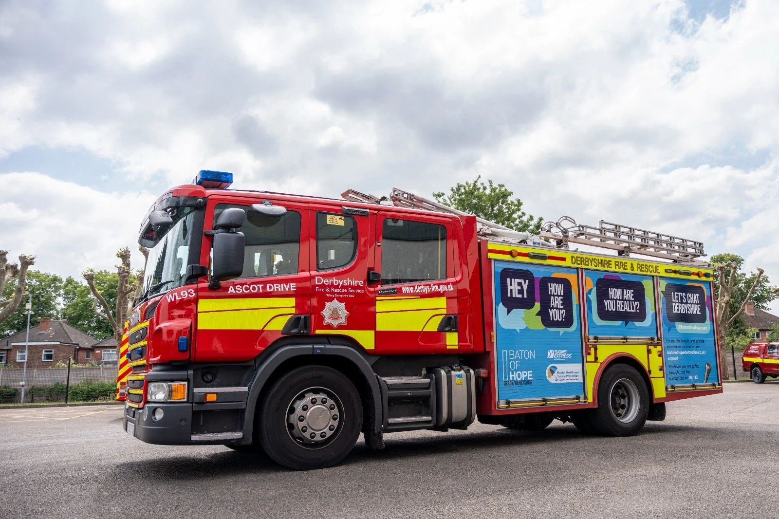 A red fire truck with yellow and blue markings, parked outdoors on a cloudy day, with trees and a fence in the background.