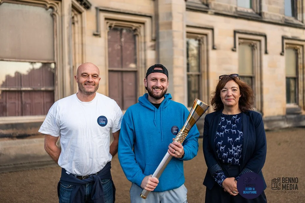 Three people outdoors in front of an old building, smiling at the camera. The person in the middle is holding a large torch. The person on the left is wearing a white T-shirt with a sticker, the person in the middle is in a blue hoodie with a cap, an