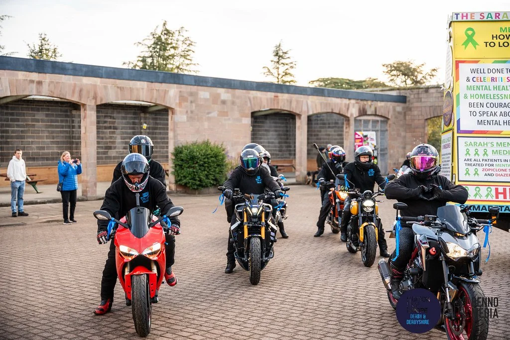 Group of motorcyclists in black jackets and helmets, riding street bikes, gathered in a parking lot near a brick wall with some trees and a colorful signboard in the background.