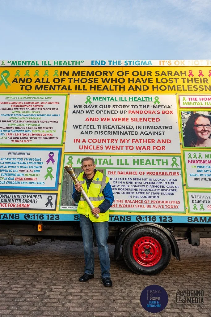 A man standing in front of a colorful informational display about mental health awareness, holding a trophy, wearing a yellow safety vest, with a truck behind him that has green and pink ribbons and various text about mental health issues.