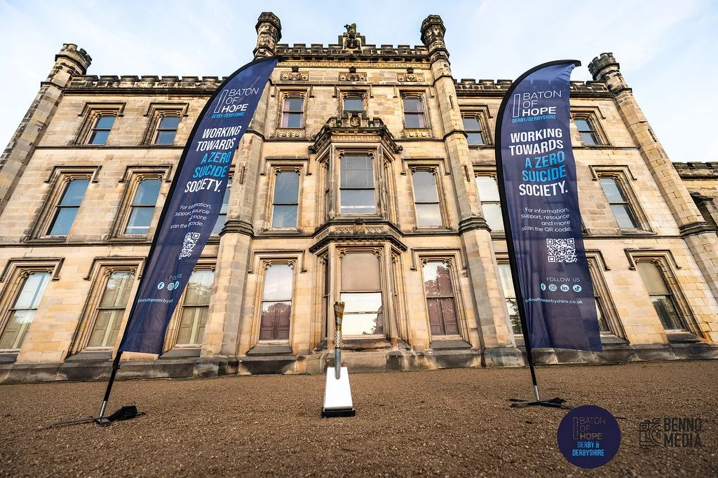 Two large blue flags on either side of a historic stone building with multiple windows, promoting a 'Baton of Hope' event supporting a zero suicide society. The flags are attached to poles and display messaging about working towards preventing suicid