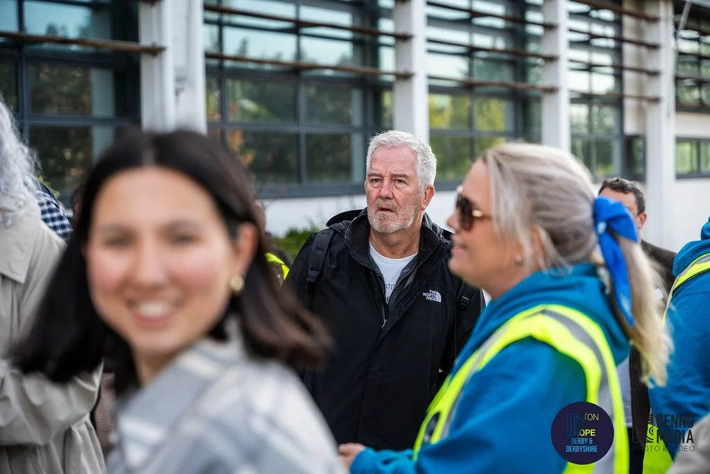 People gathered outdoors near a modern building with glass windows, including a man with white hair and a black jacket, a woman with gray hair and sunglasses wearing a safety vest, and a smiling woman in the foreground.