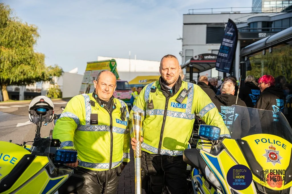 Two police officers in fluorescent yellow jackets stand in front of police motorcycles, one holding a torch, during a public event with people in the background.