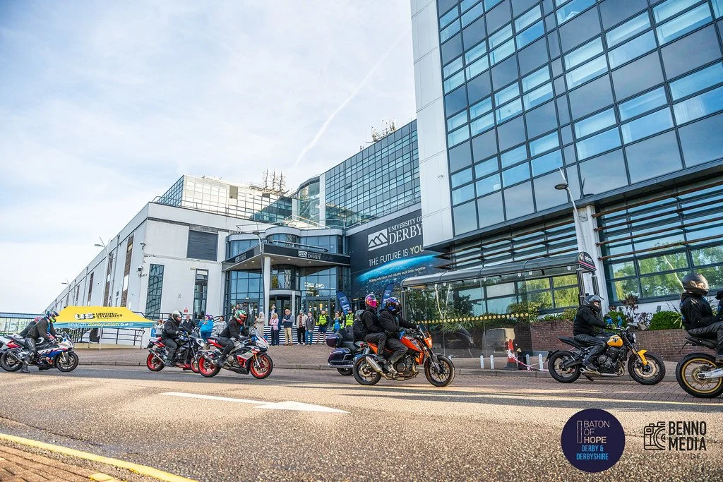 People riding motorcycles in front of the University of Derby building on a city street.