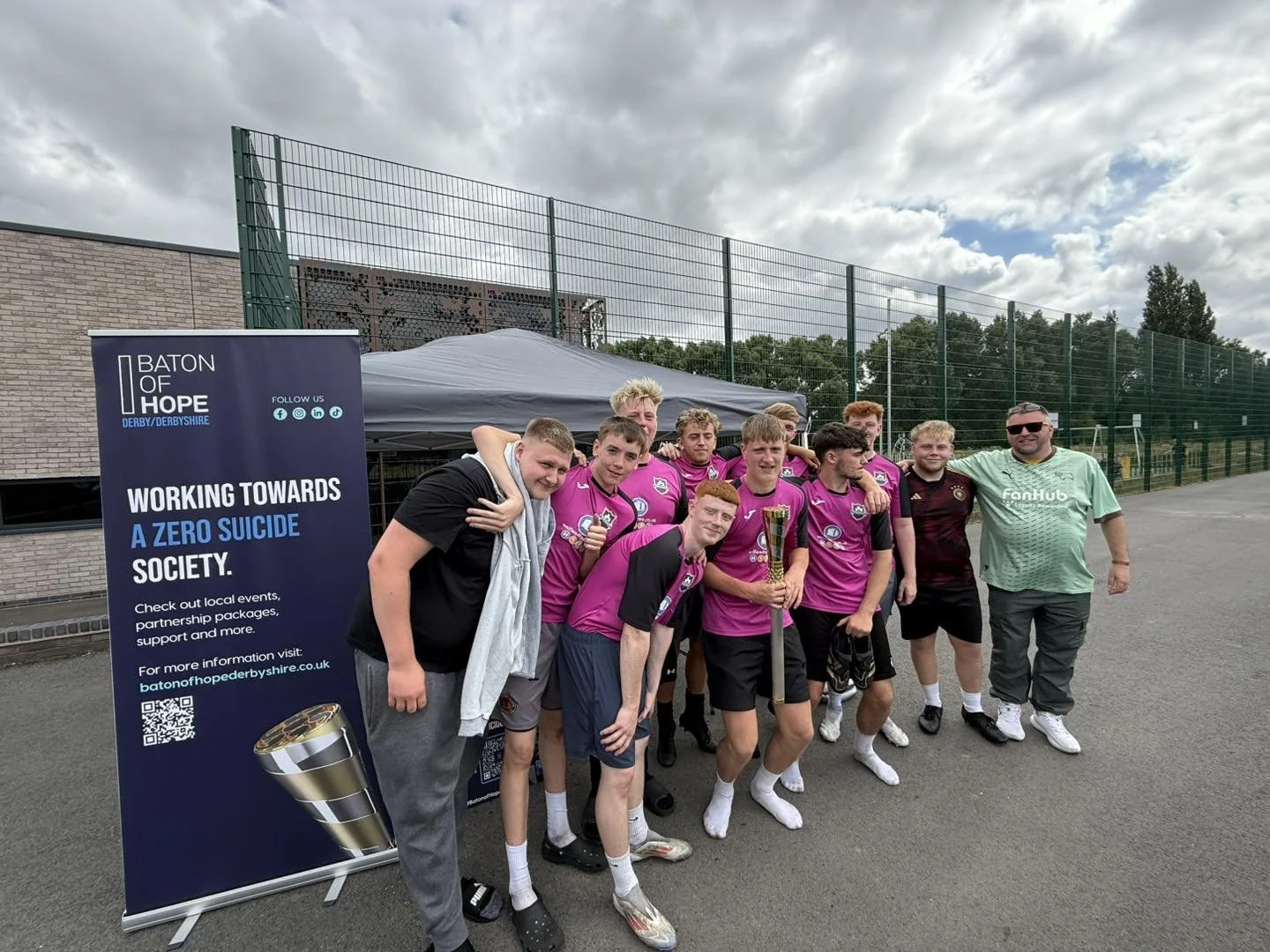 Group of young soccer players in pink jerseys celebrating with a trophy, standing next to a man in sunglasses and a green shirt, outdoor sports field in the background.