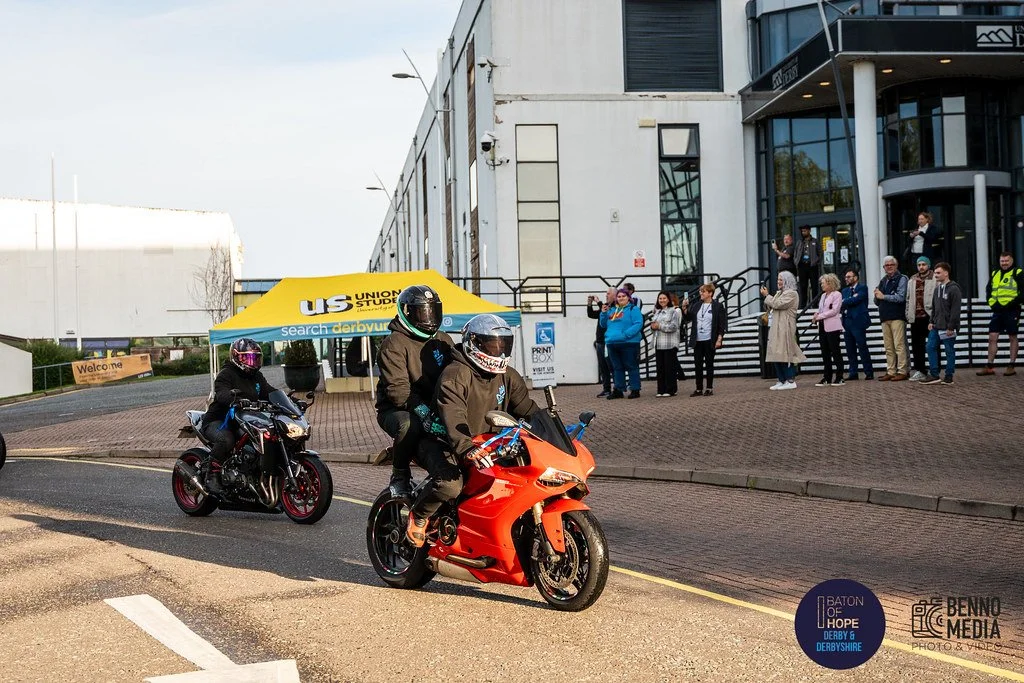 Two motorcyclists riding on the street, with a group of people waiting outside a modern building. There is a yellow tent with a blue sign in the background, and some people are taking photos or videos.