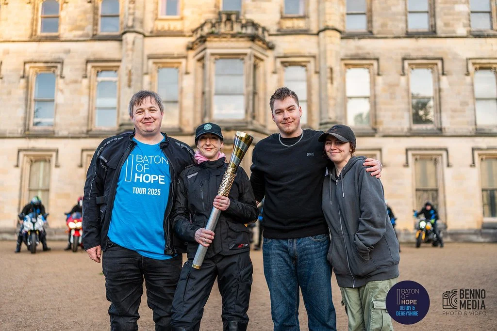 Four people standing in front of a historic building, smiling, with a group of motorcyclists in the background. One person holds a torch, and there is a logo at the bottom right corner that reads 'Baton of Hope Derby & Derbyshire'.