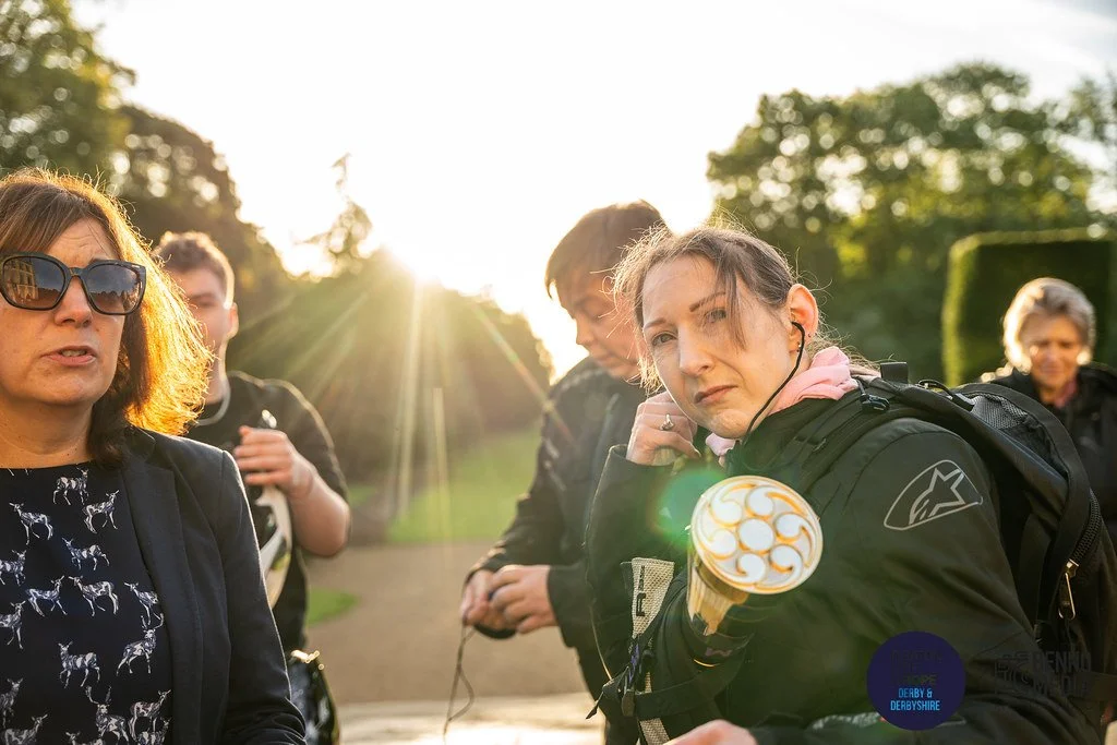 Group of women and a young man outdoors during sunset, some wearing motorcycle gear, one woman wearing sunglasses, in a park with trees.