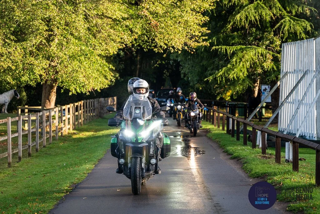 A group of motorcyclists riding on a narrow paved path surrounded by green trees and grass, with a white fence on the right side and a white horse behind the fence on the left.